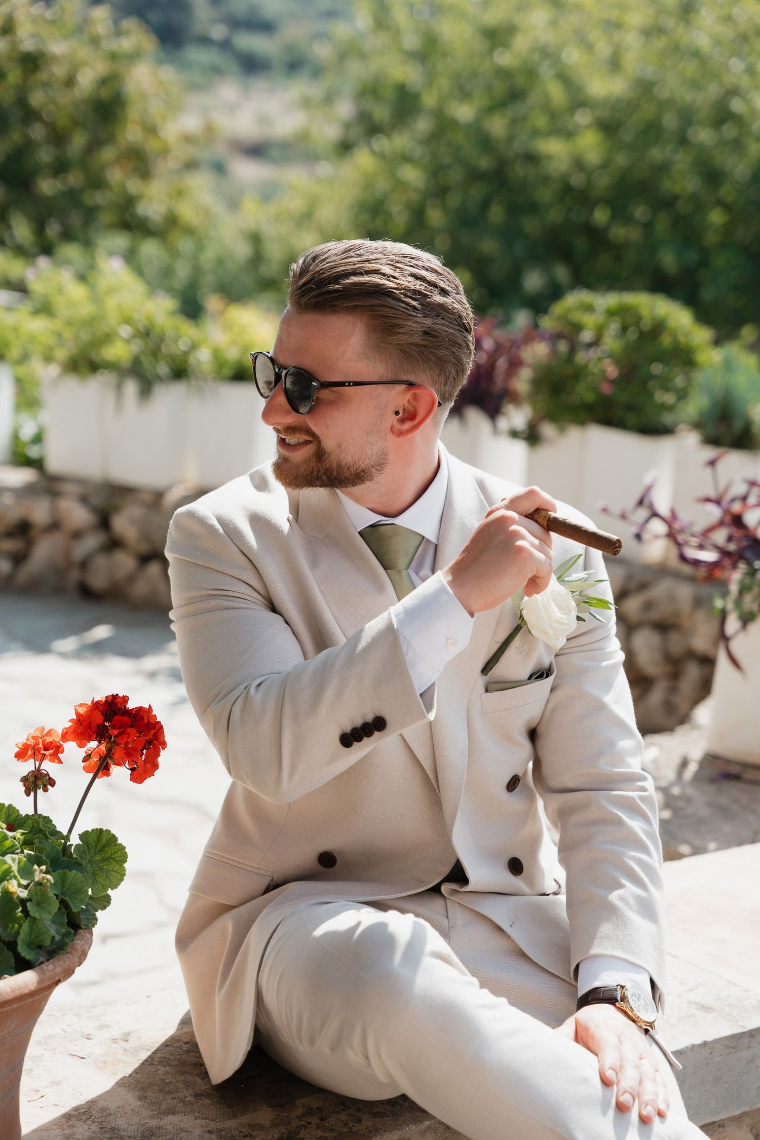 A person in a white suit and sunglasses relaxes outdoors on a sunny terrace with red flowers in the background.