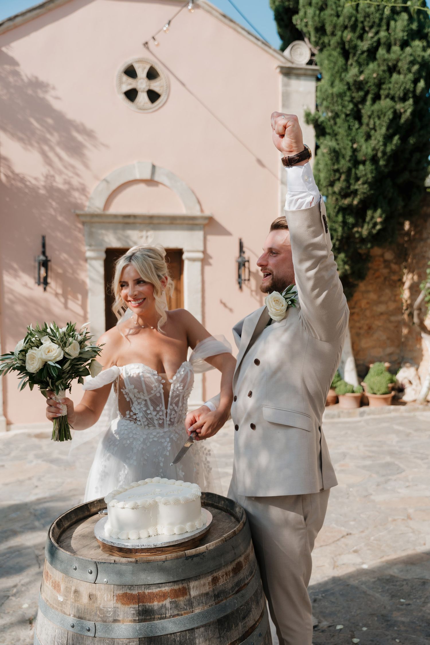 Newlyweds celebrate joyfully next to a wine barrel outside a pink Mediterranean church on their wedding day.
