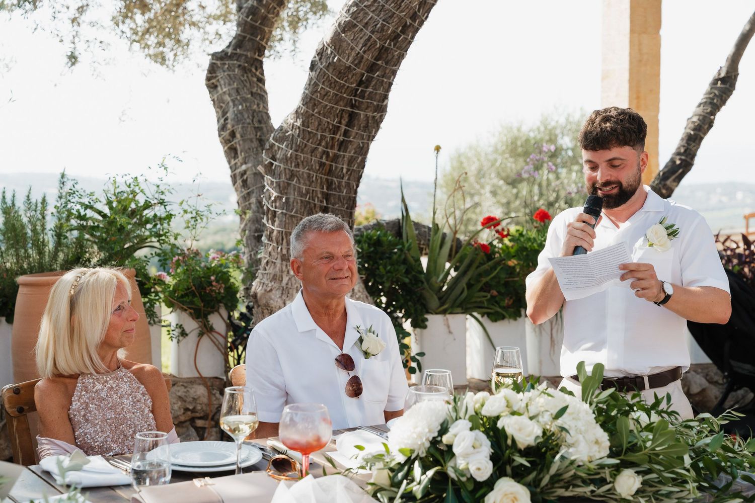Wedding reception guests seated at an outdoor table with white floral centerpieces while someone gives a speech.