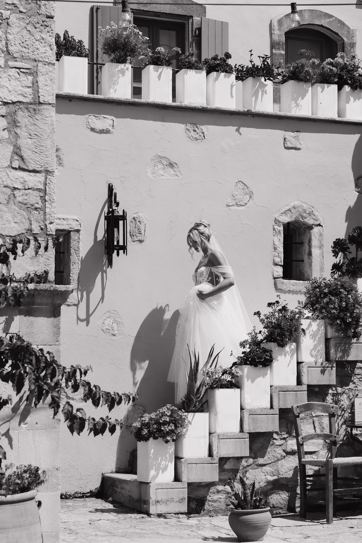 A black and white photograph shows a figure in a flowing dress ascending stone steps adorned with potted plants and flowers.