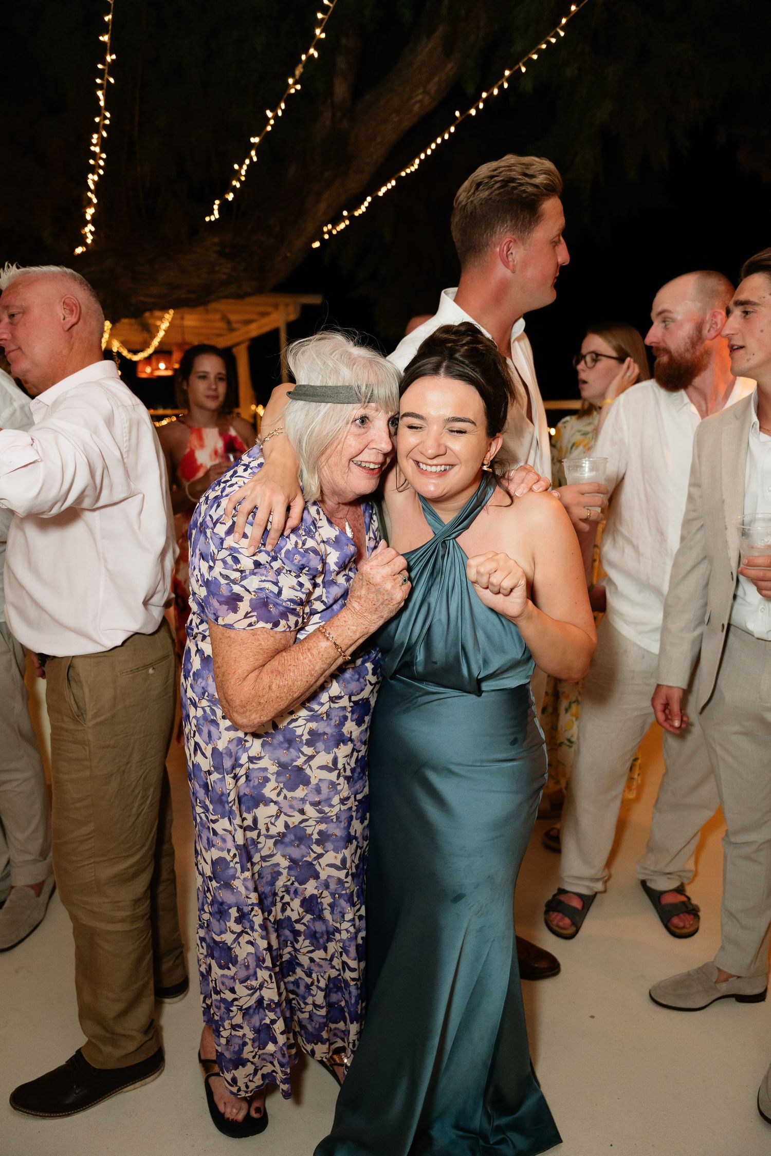 Two guests share a joyful moment at a wedding reception, one wearing a floral dress and another in a teal halter gown.