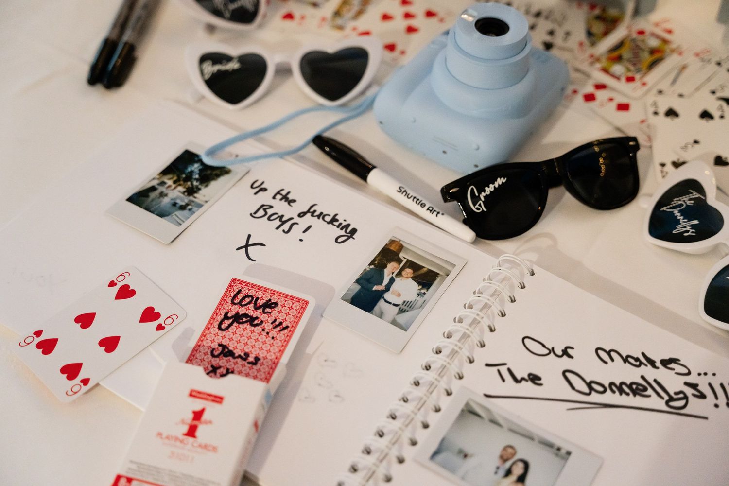 Playing cards, heart-shaped sunglasses, and a light blue instant camera arranged with photos and notebook on white surface.