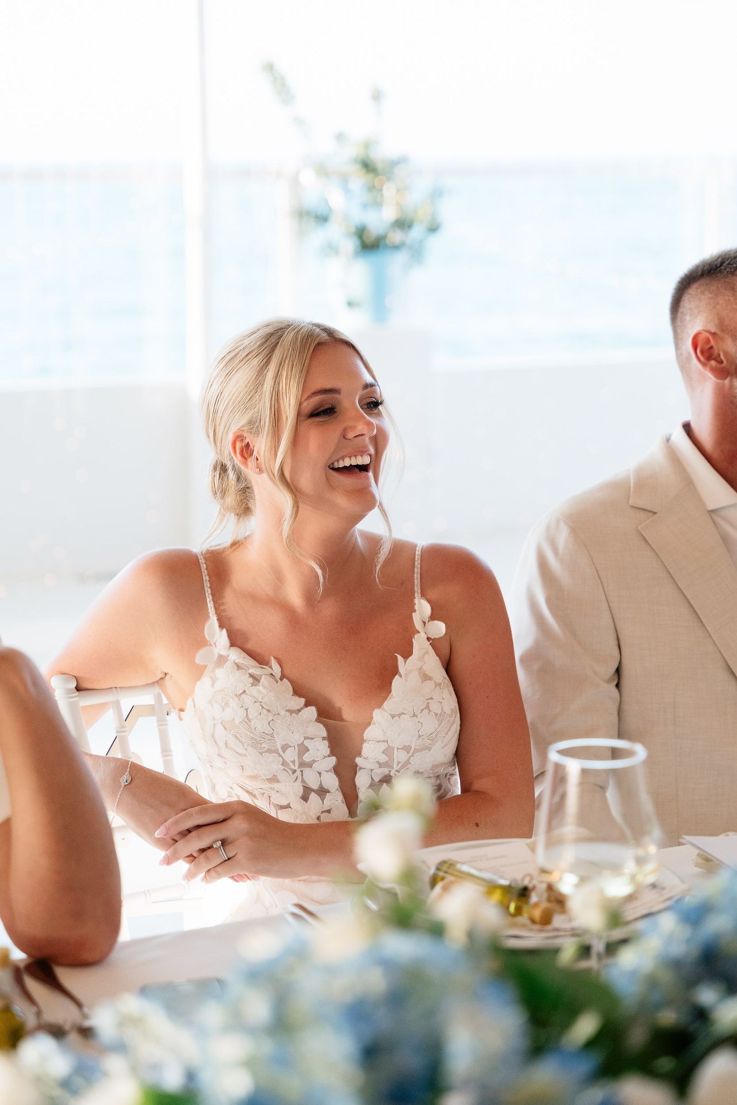 A person in a white wedding dress laughs joyfully while seated at an elegant reception table with floral decor.