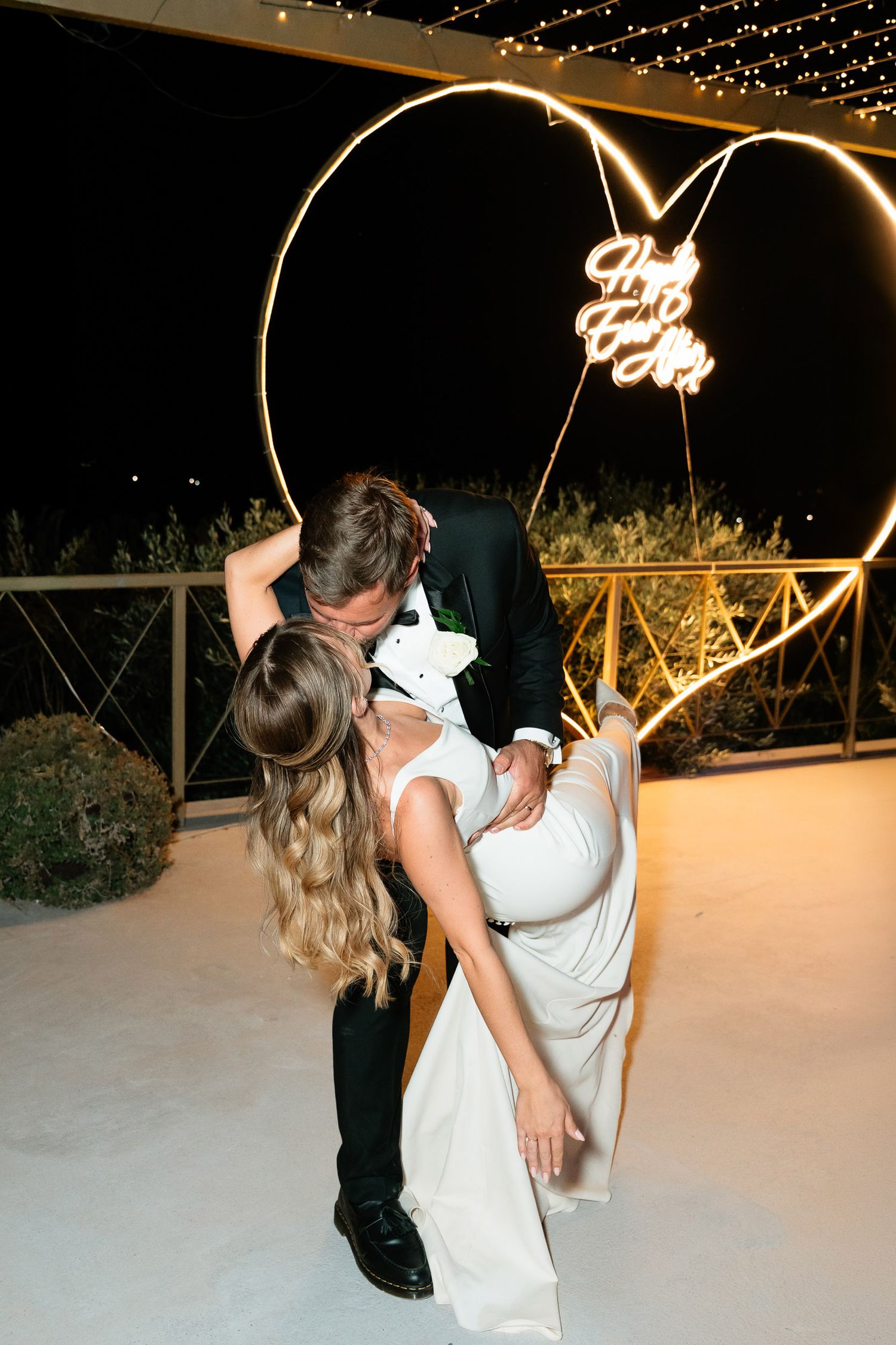 A romantic couple dancing under twinkling string lights at an outdoor venue in the evening.