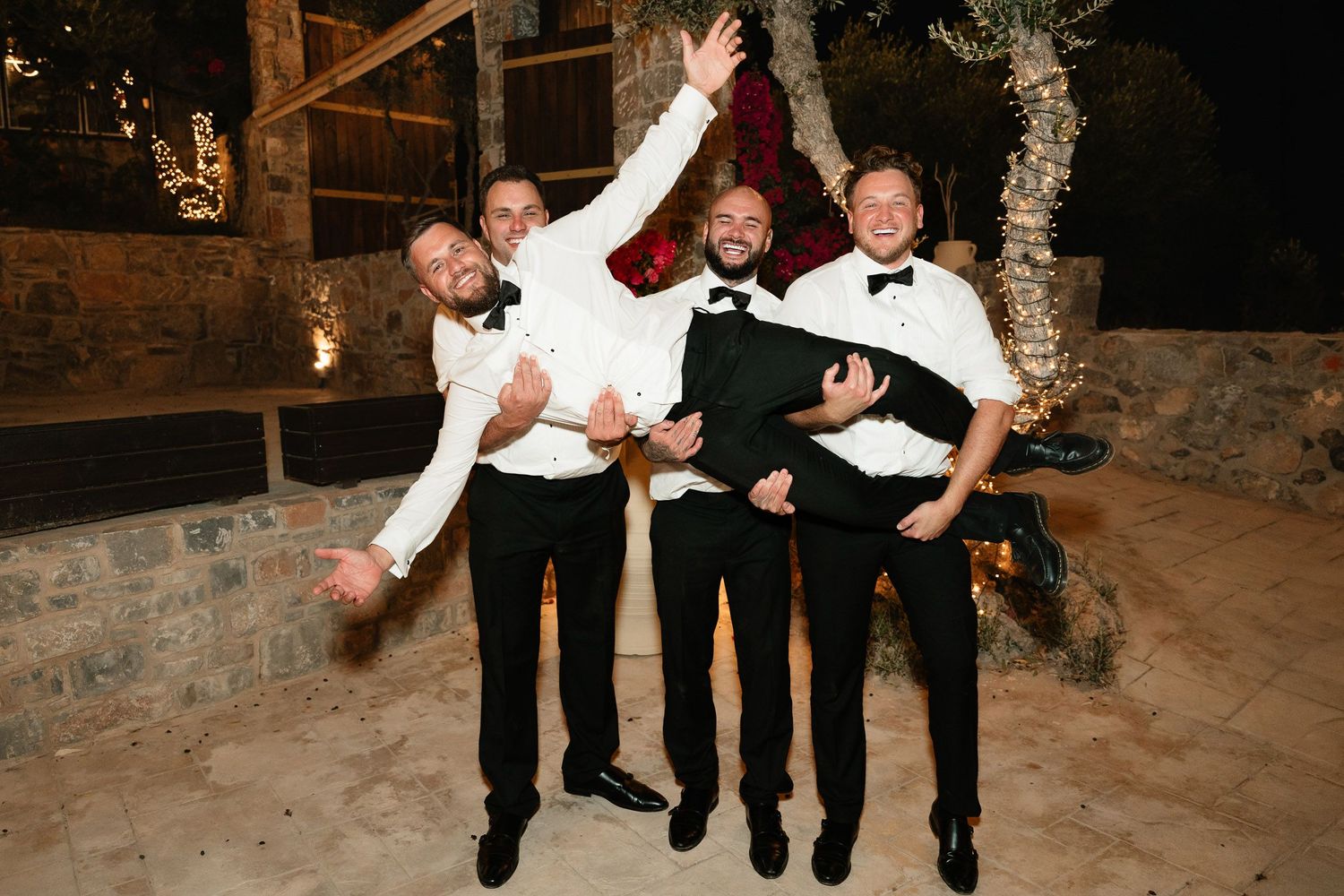 Wedding party members in black tuxedos and bow ties strike playful poses during an outdoor evening celebration.