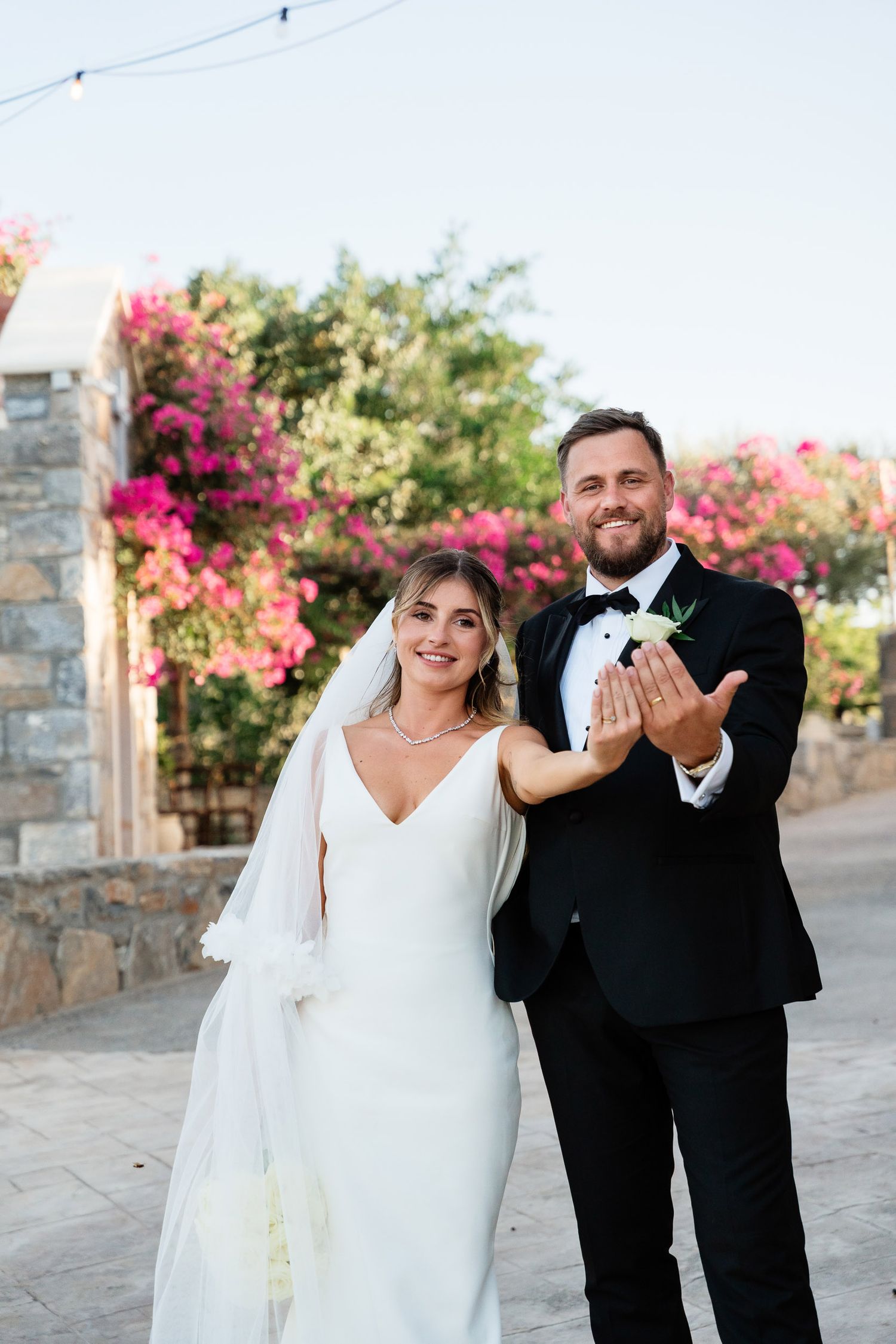 A couple in wedding attire share joyful candid moments in front of pink bougainvillea flowers on a stone walkway.