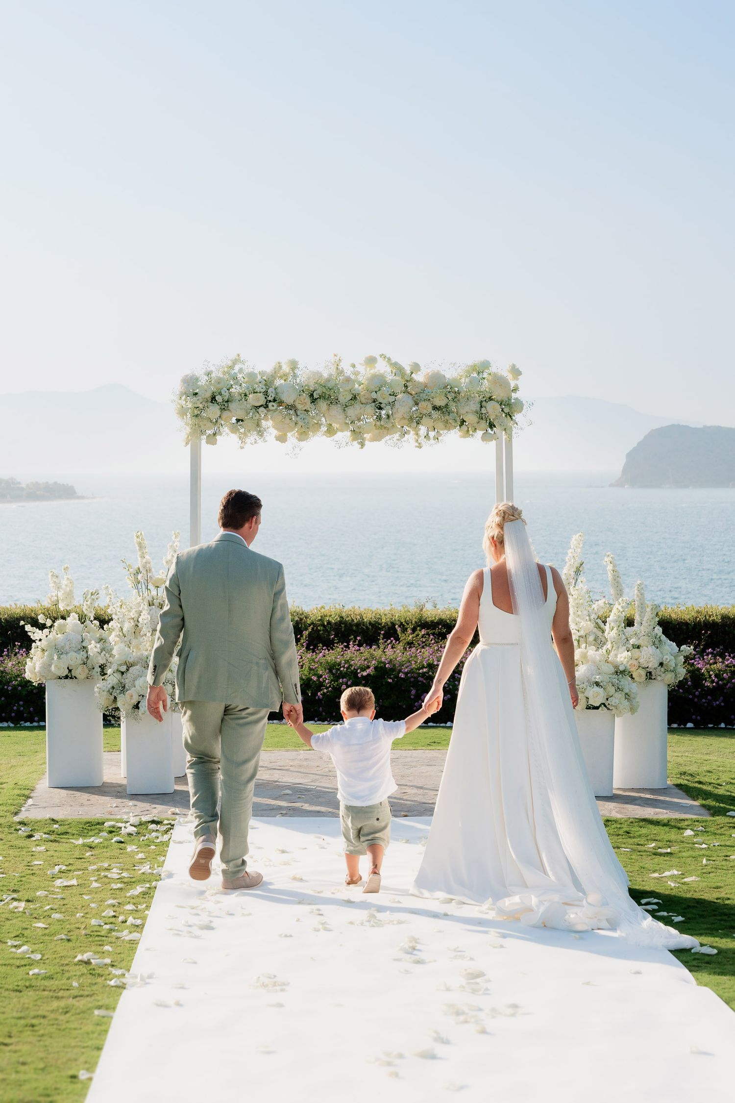 A wedding ceremony setup on a scenic oceanfront lawn with white floral arrangements and a flower-adorned arch.