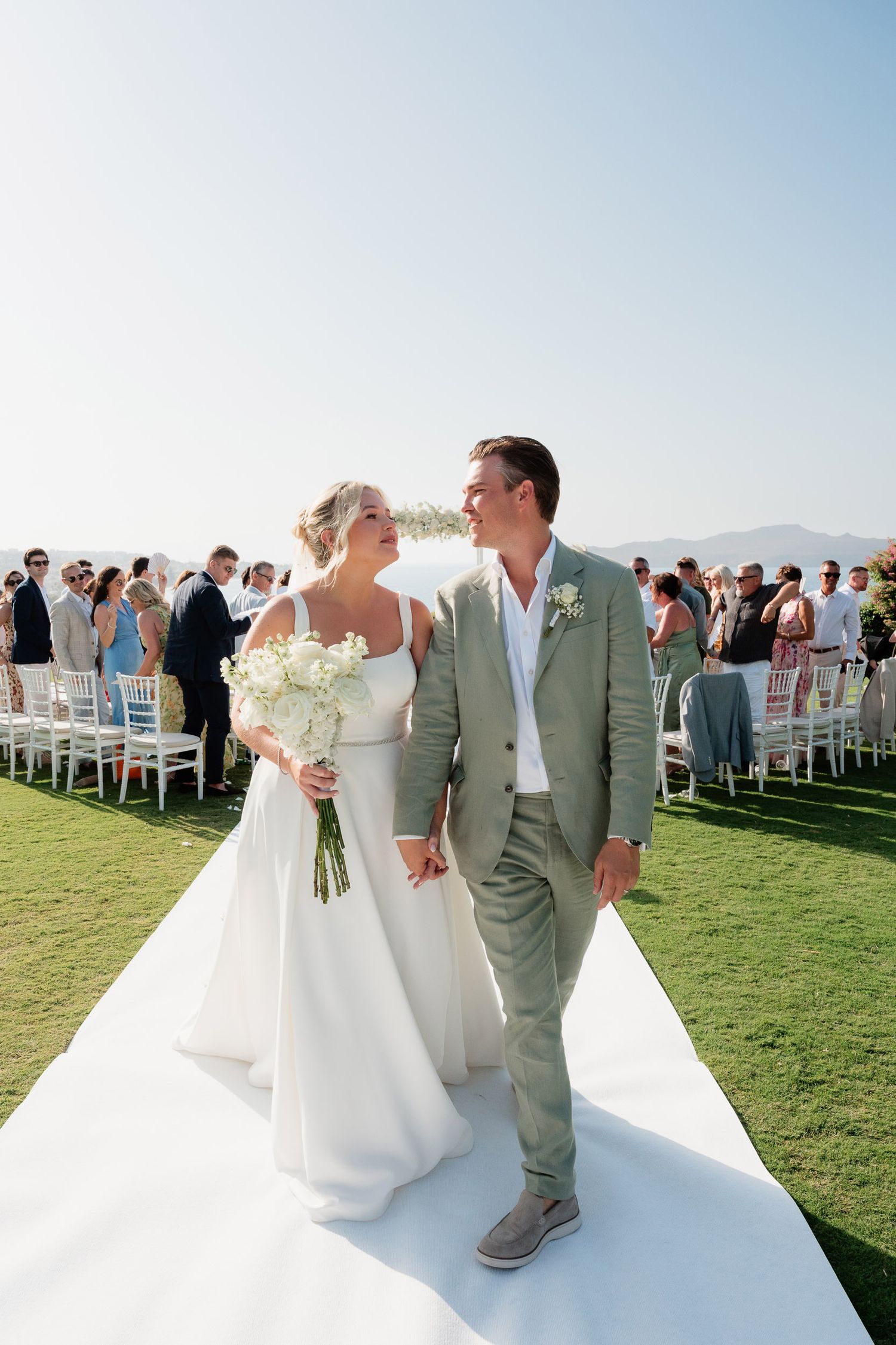 A couple shares their first dance at an outdoor wedding ceremony on a grassy lawn with guests watching in the background.
