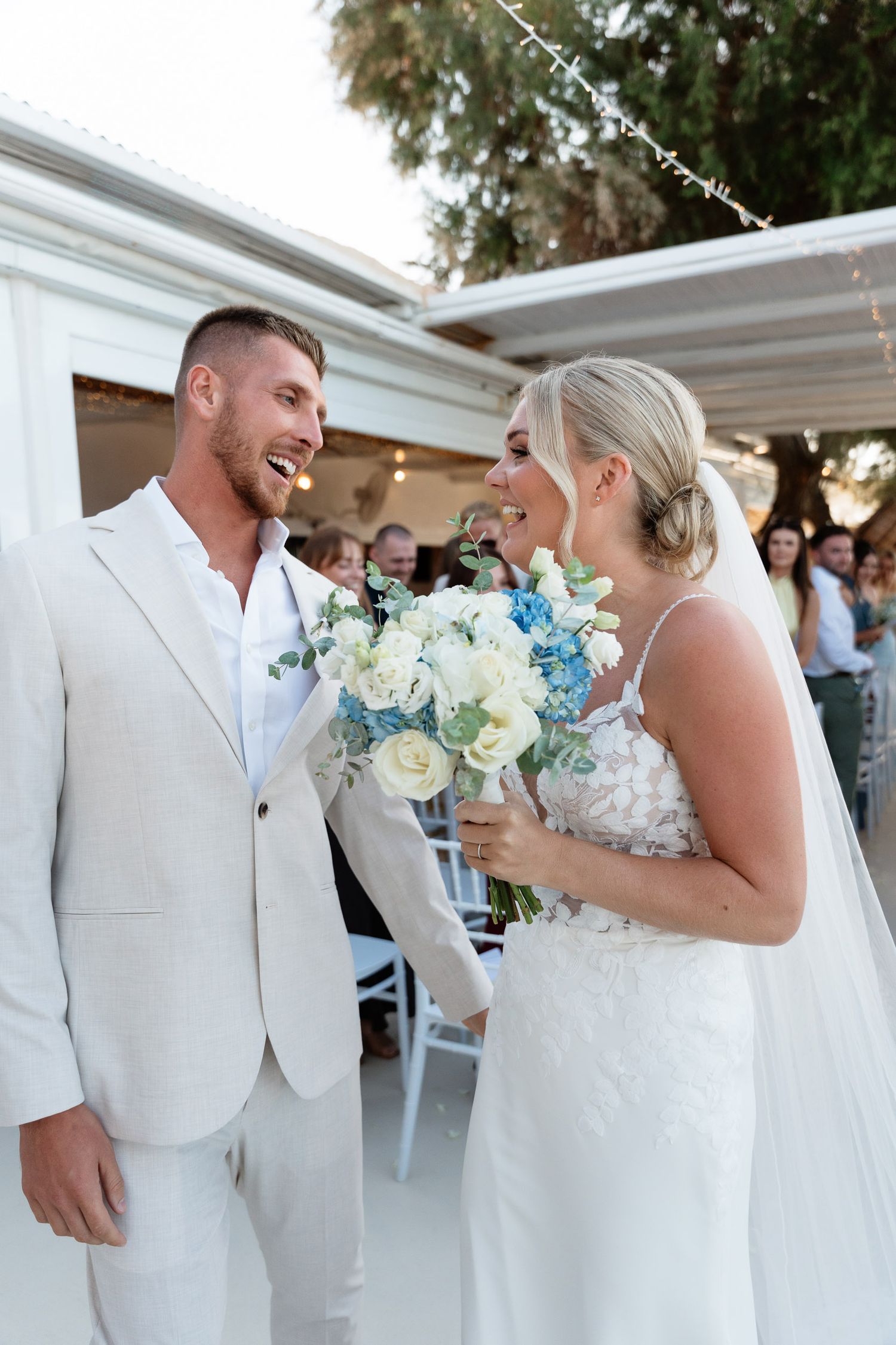 Joyful wedding couple shares a candid moment during their outdoor ceremony with white and blue floral bouquet details.