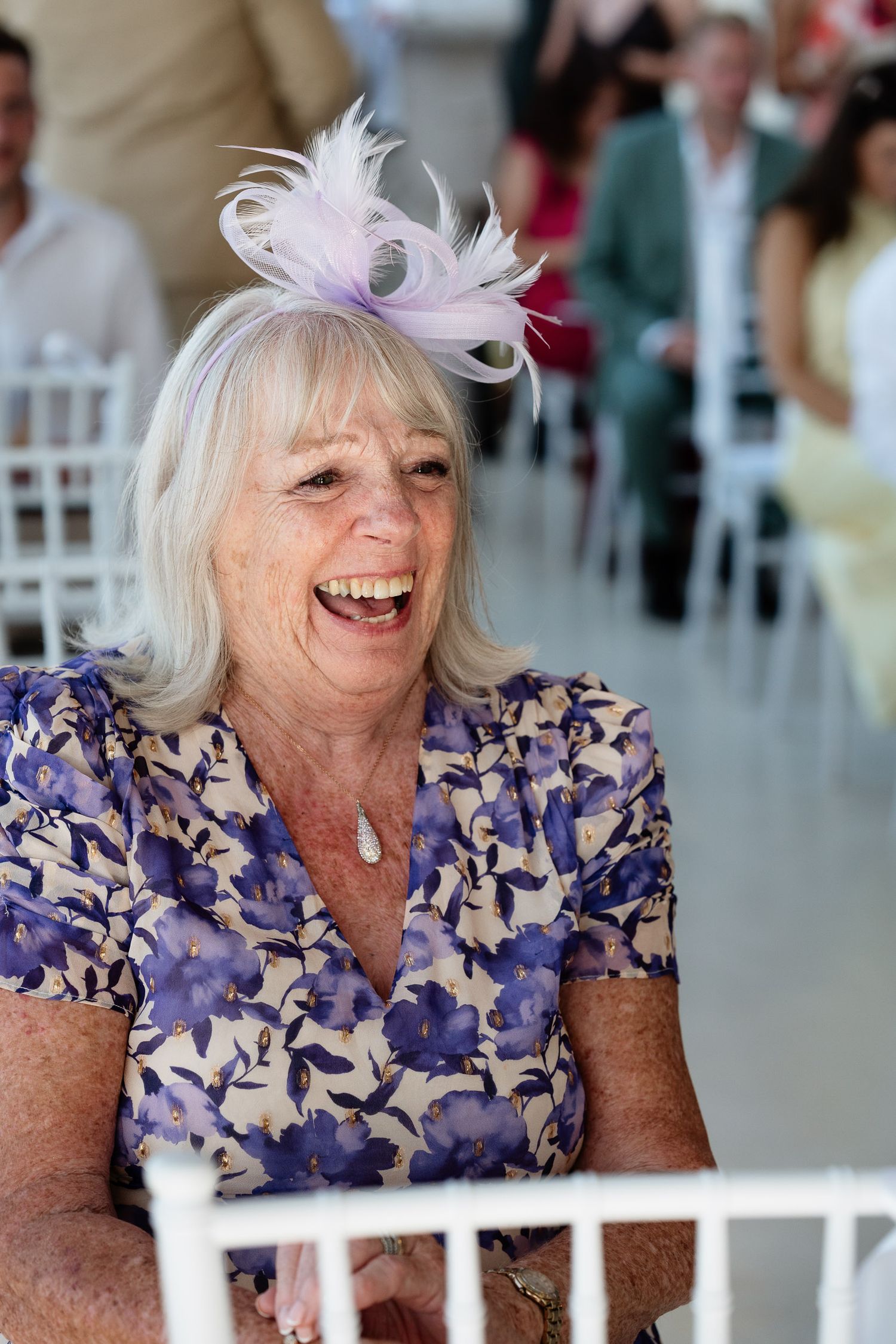 A person in a blue floral dress laughs joyfully while seated at what appears to be a social gathering.