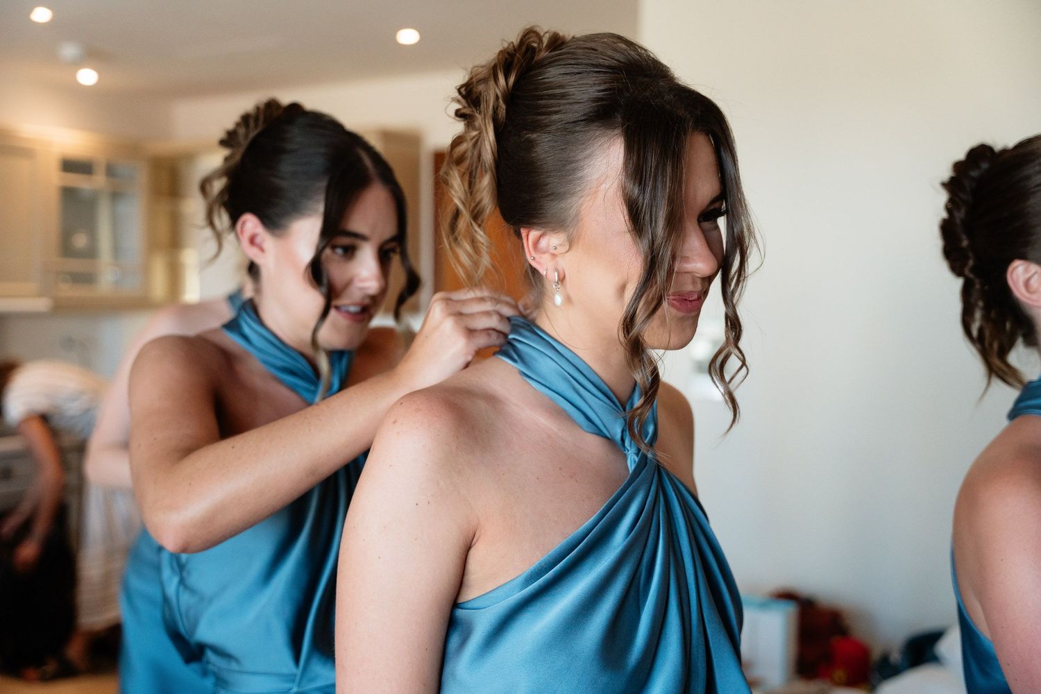 Bridesmaids in turquoise halter dresses help each other get ready before a wedding ceremony.