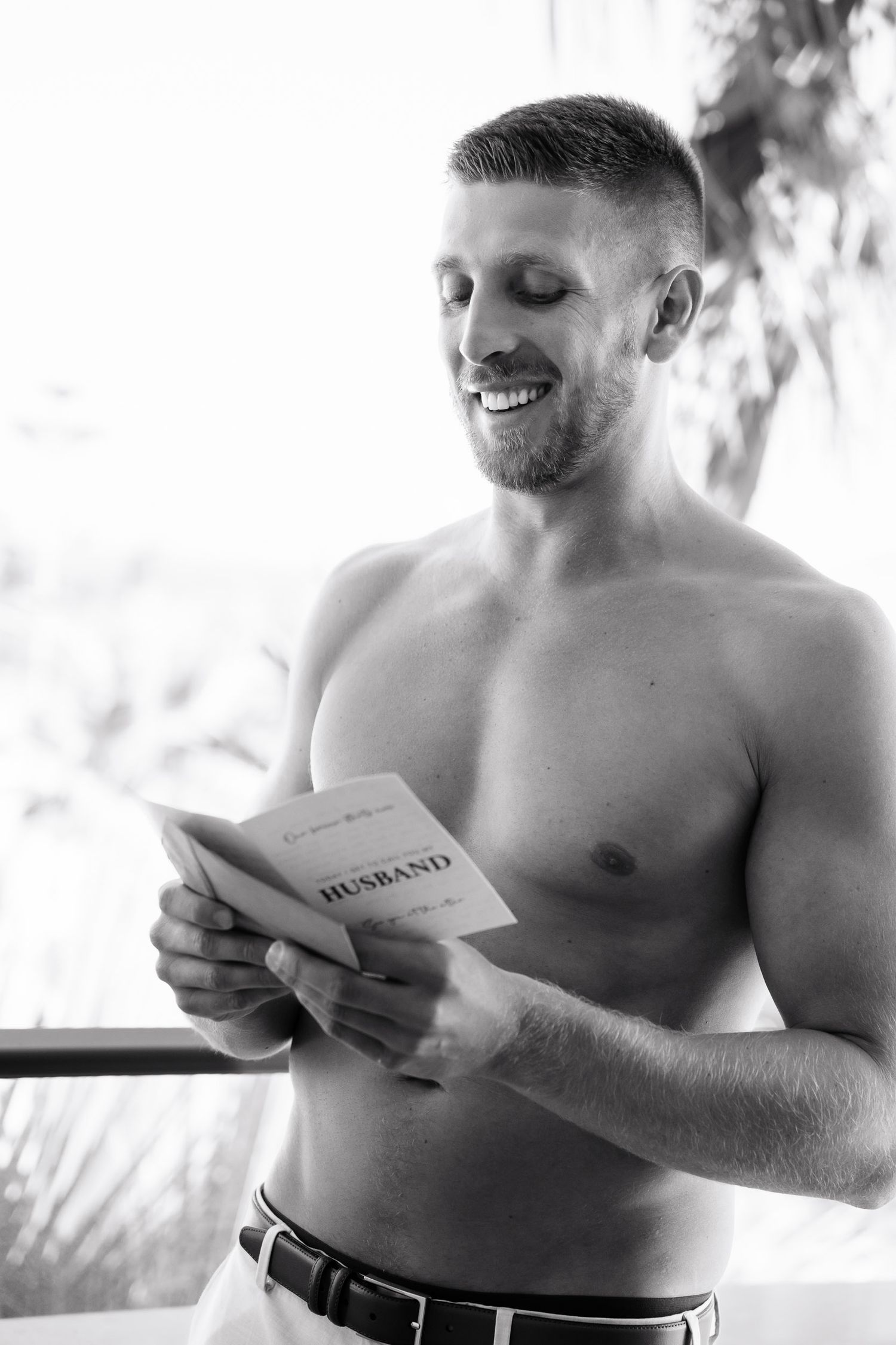 Black and white photo of shirtless person reading a letter or note with a joyful expression.