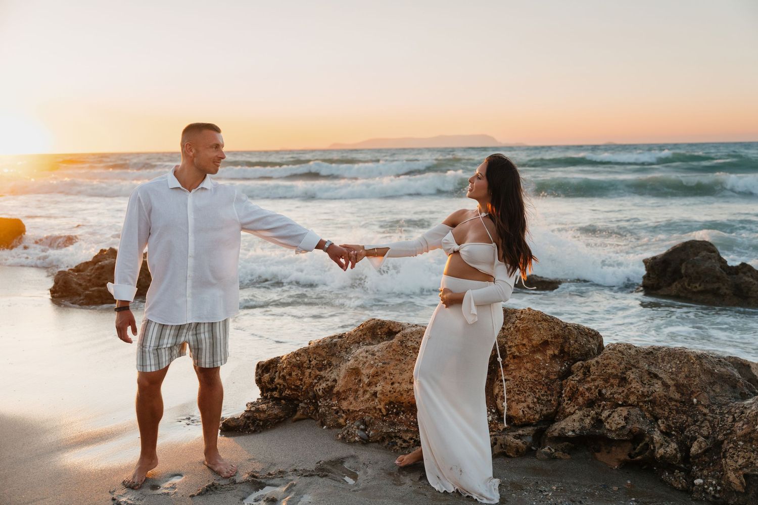 A couple in white outfits holds hands on a rocky beach during golden hour with ocean waves in the background.