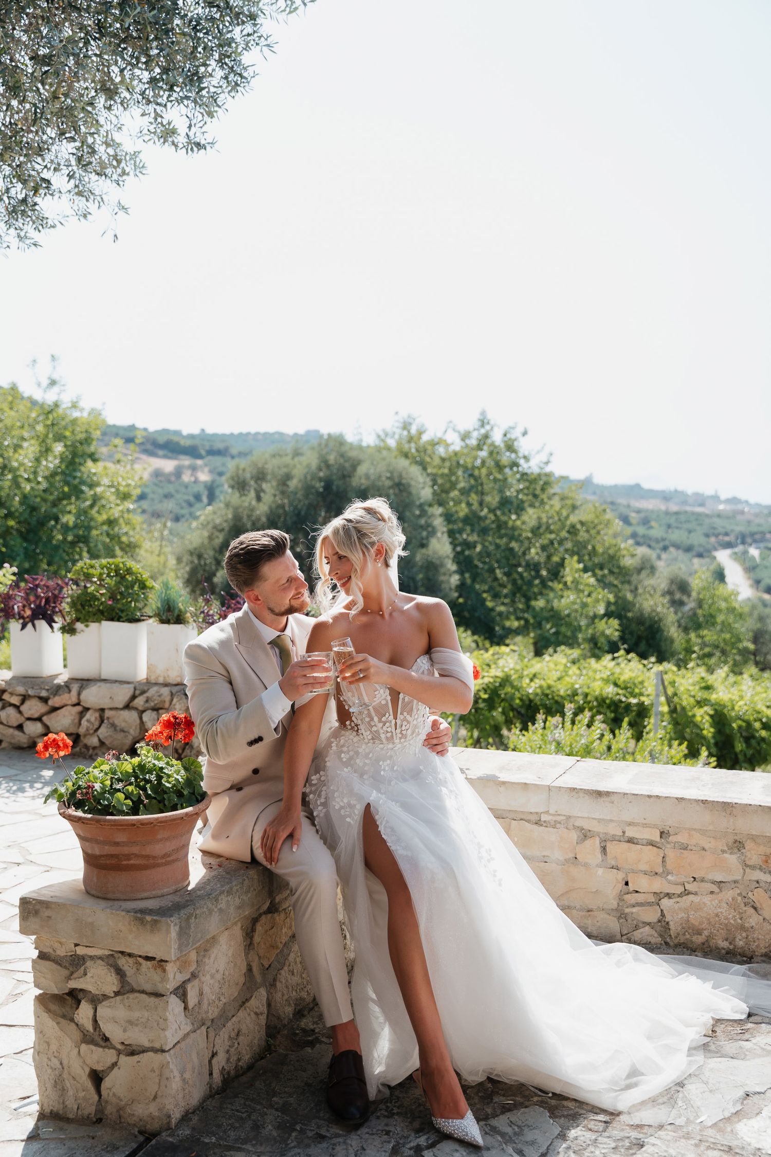 A couple in wedding attire sits together on a stone wall overlooking a scenic Tuscan countryside on a sunny summer day.