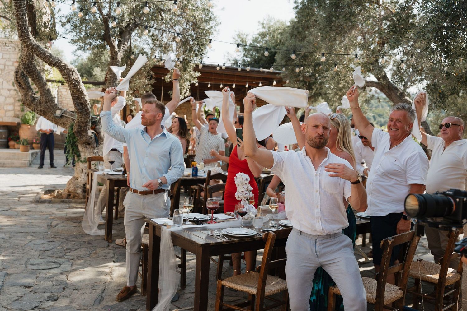 Outdoor wedding reception guests enjoying a casual celebration under trees with rustic wooden tables and chairs.