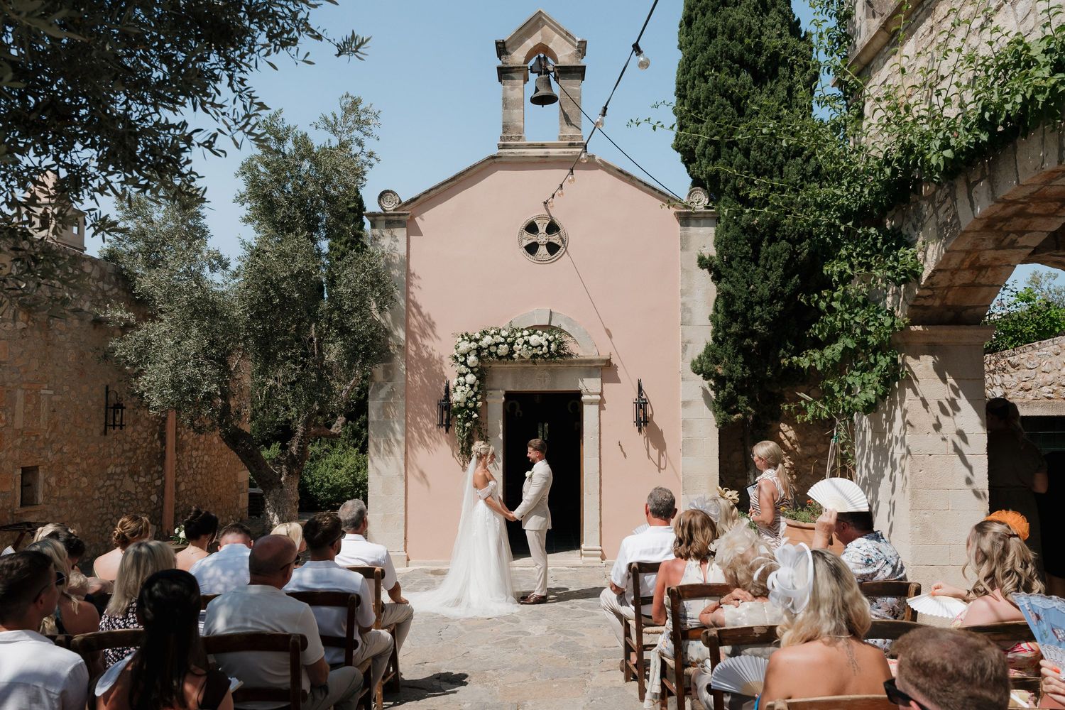 A collection of small pink and beige chapel buildings with guests gathering outside for a wedding celebration.