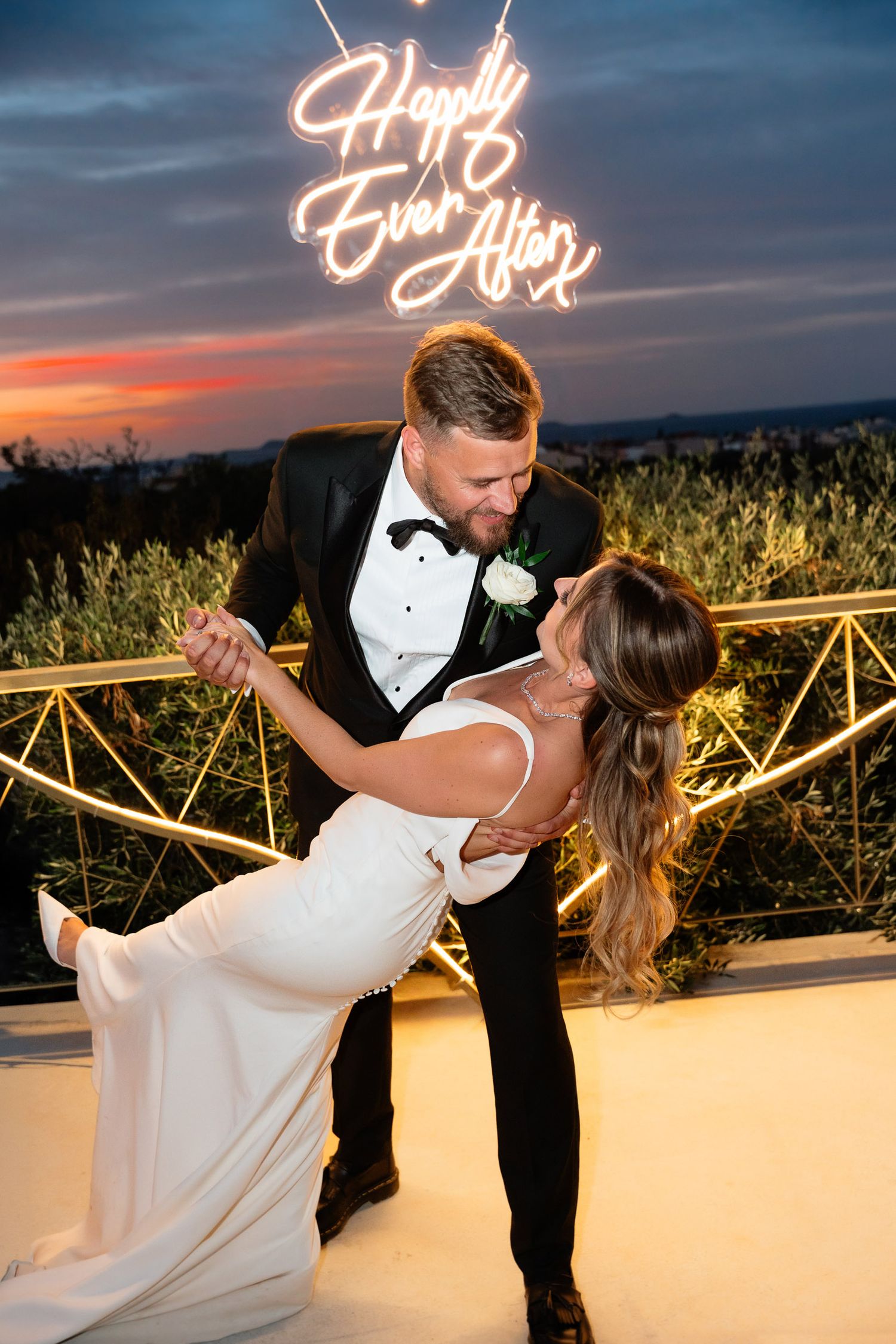 A romantic wedding dance on a terrace at sunset with a glowing neon sign overhead.