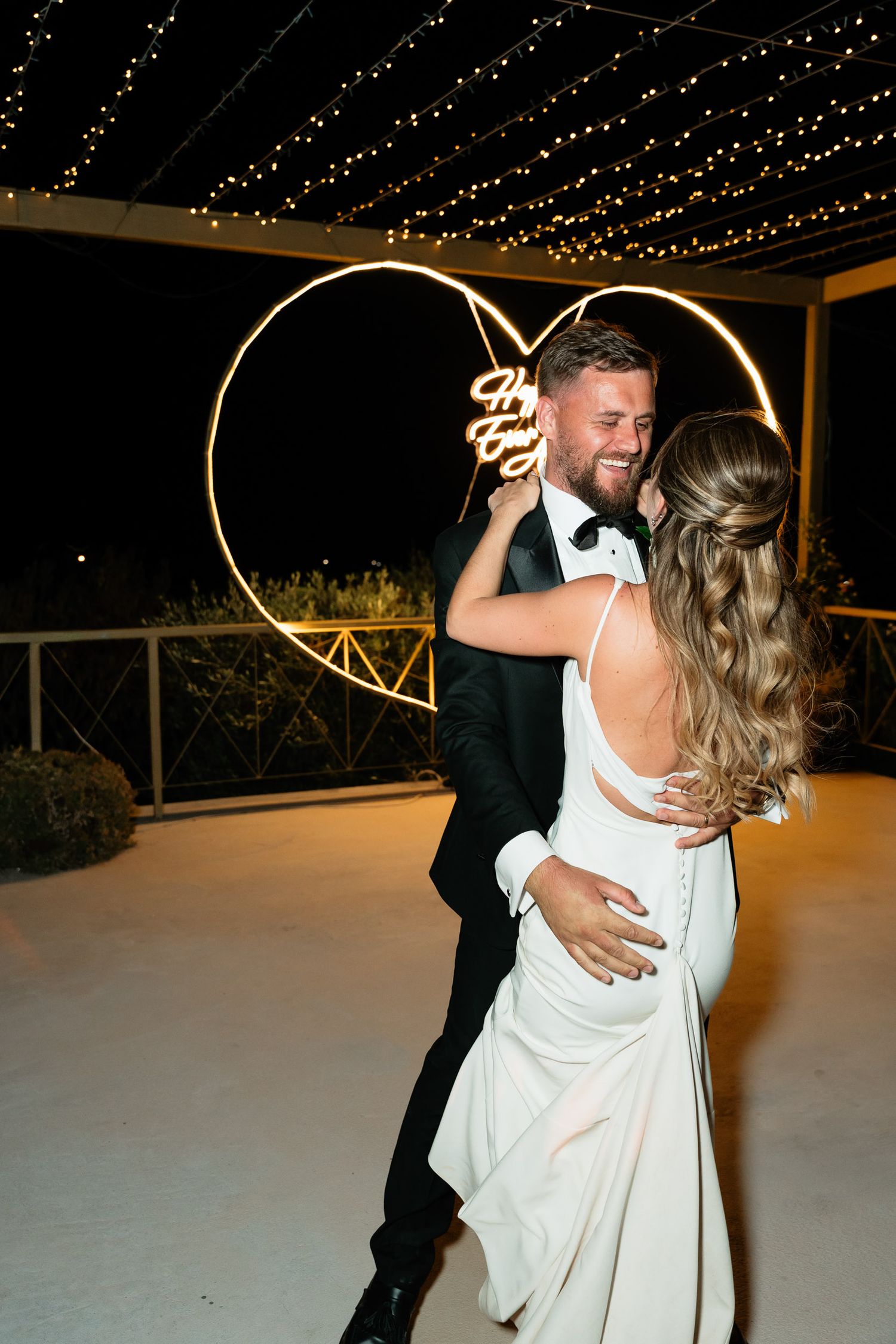 A couple dances together under twinkling string lights at an evening wedding reception, the bride wearing a flowing white dress.