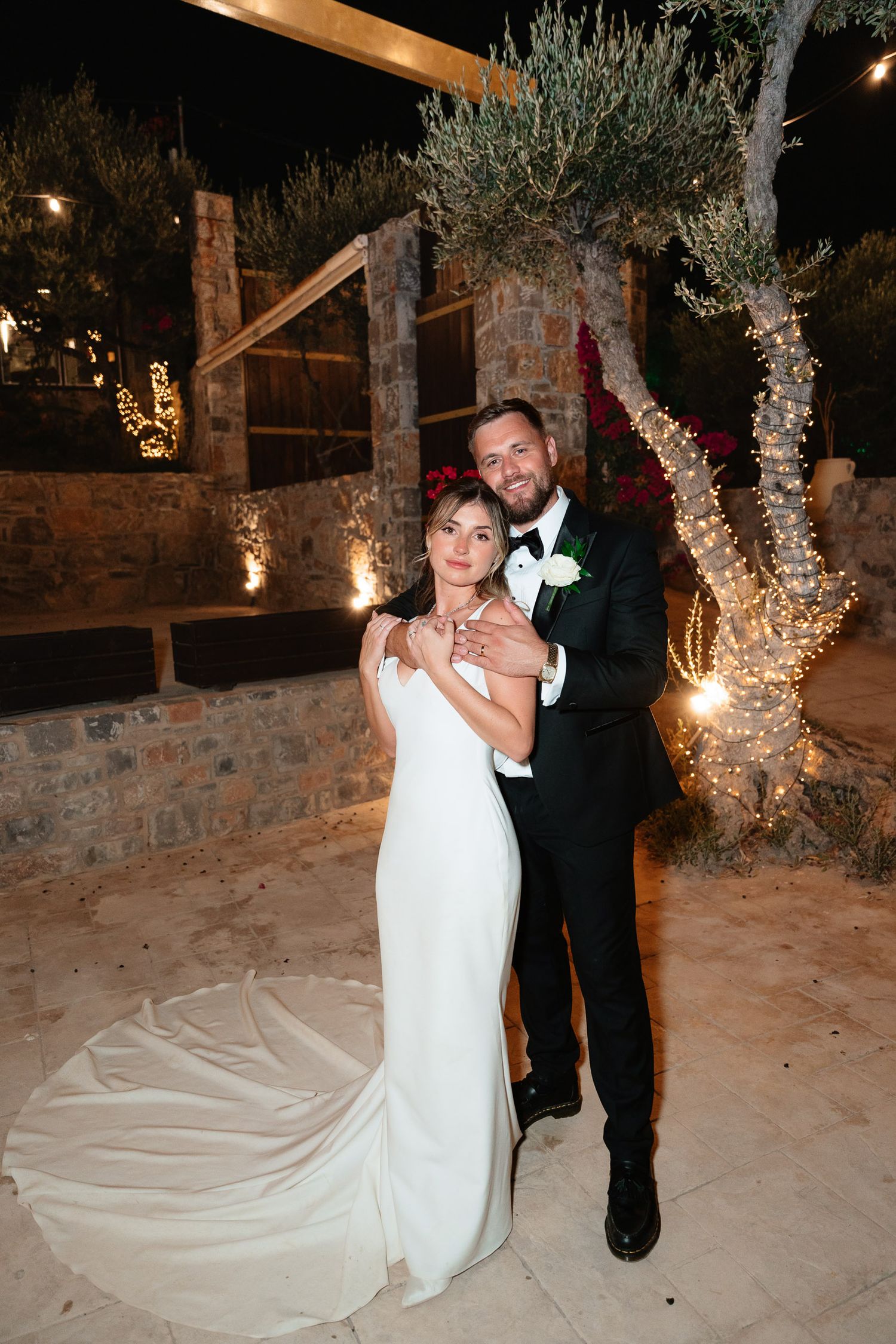 A couple dances together at night in an outdoor venue with stone walls and olive trees illuminated by warm lighting.