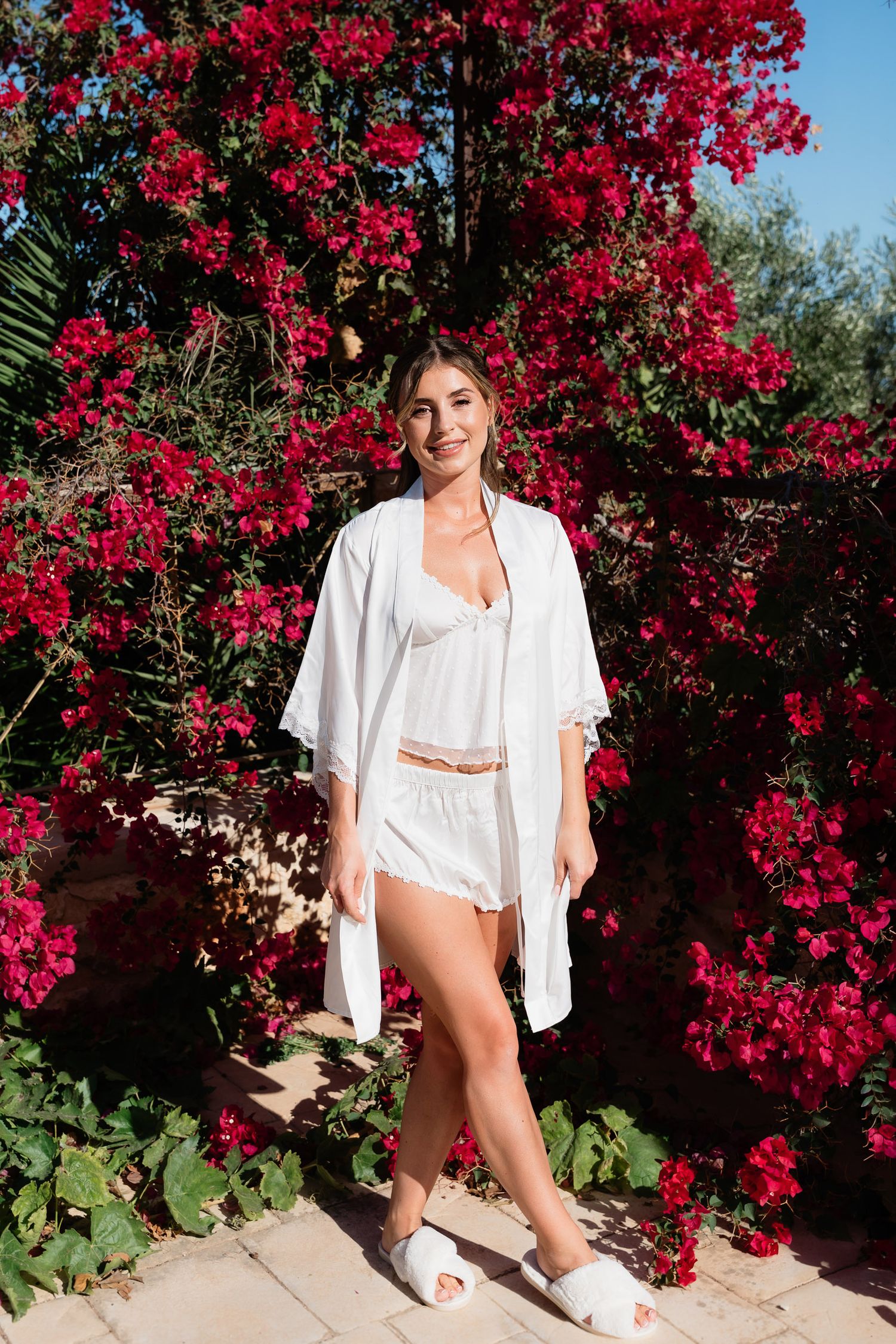 A figure in a white shirt poses against vibrant red bougainvillea flowers in an outdoor garden setting.