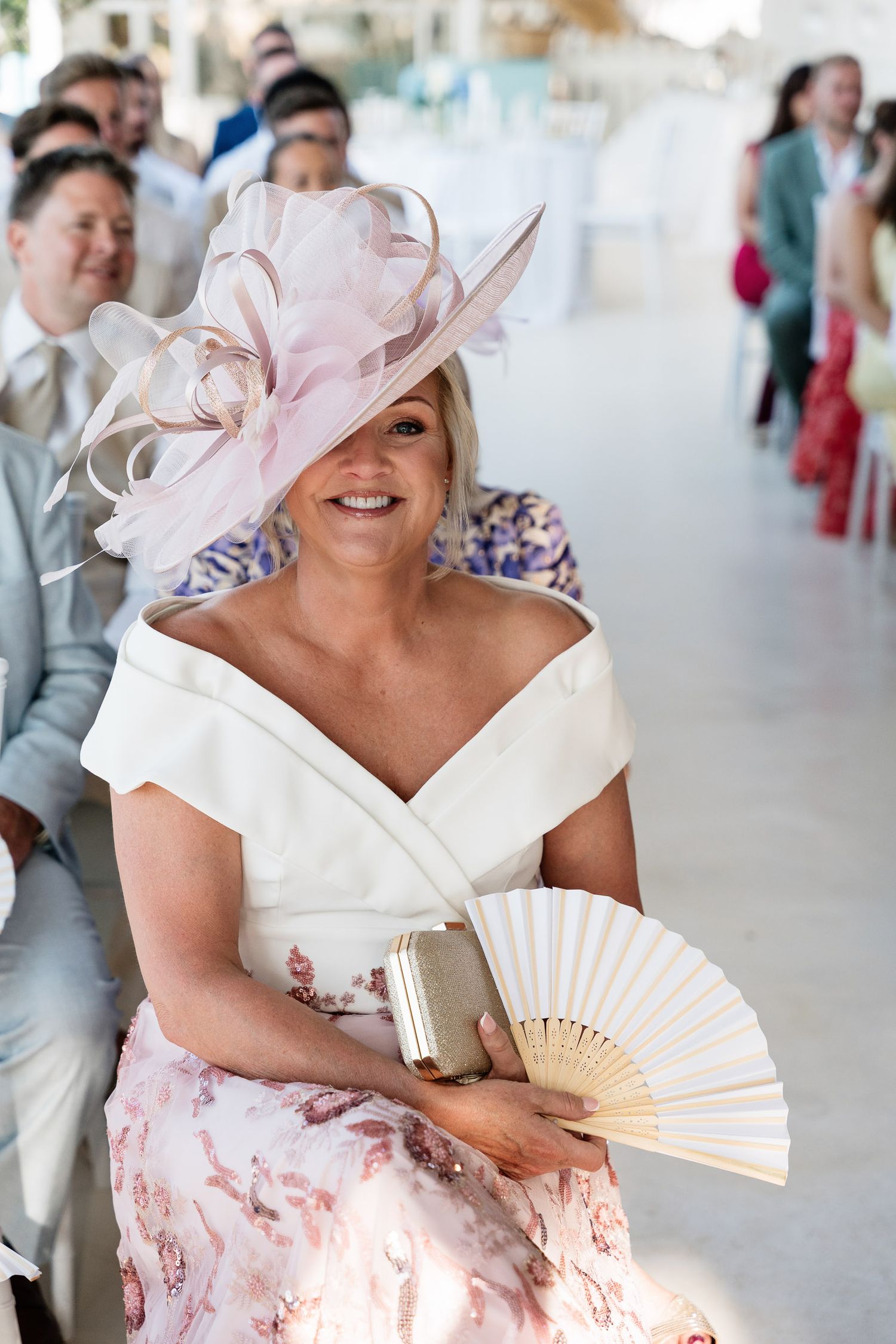 A person in a white and pink dress holds a decorative hand fan while wearing an elegant light pink hat at an event.