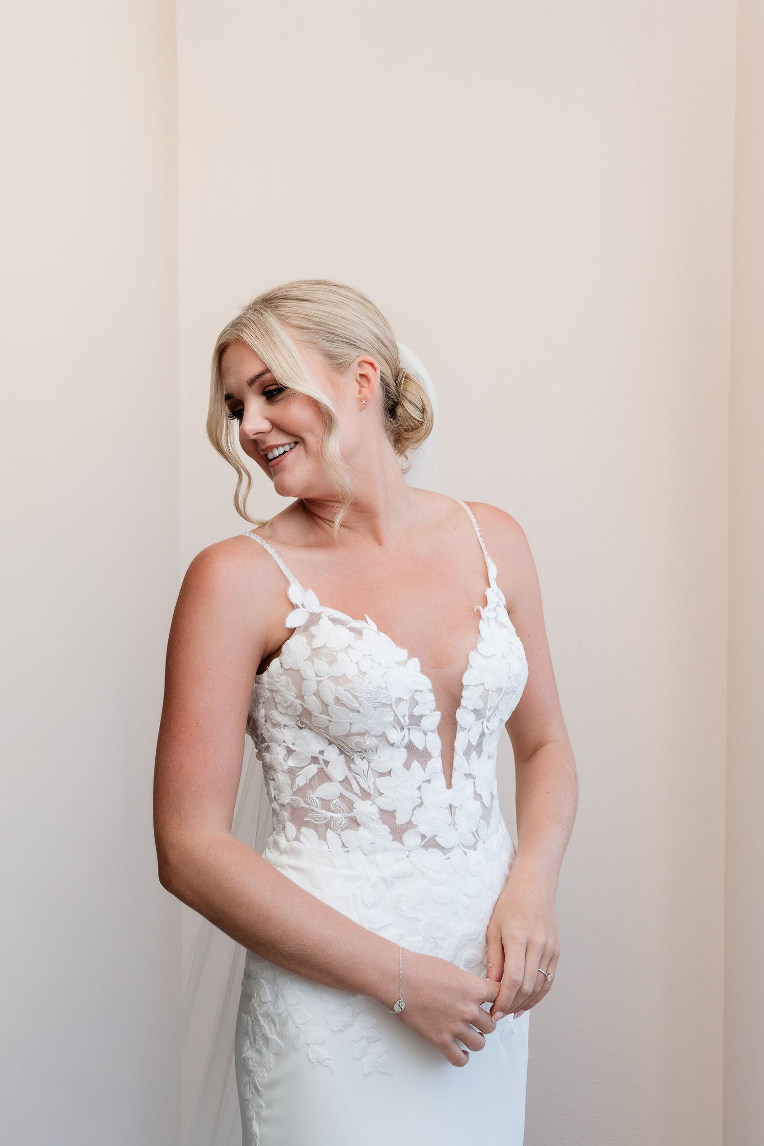 A bride in a white lace wedding dress with spaghetti straps poses against a light colored wall in three similar poses.
