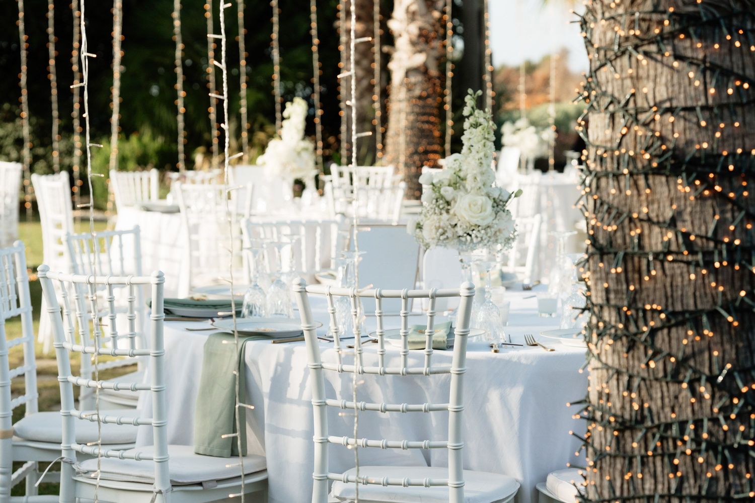 Elegant outdoor wedding reception setup with white chiavari chairs and palm trees in a tropical garden setting.