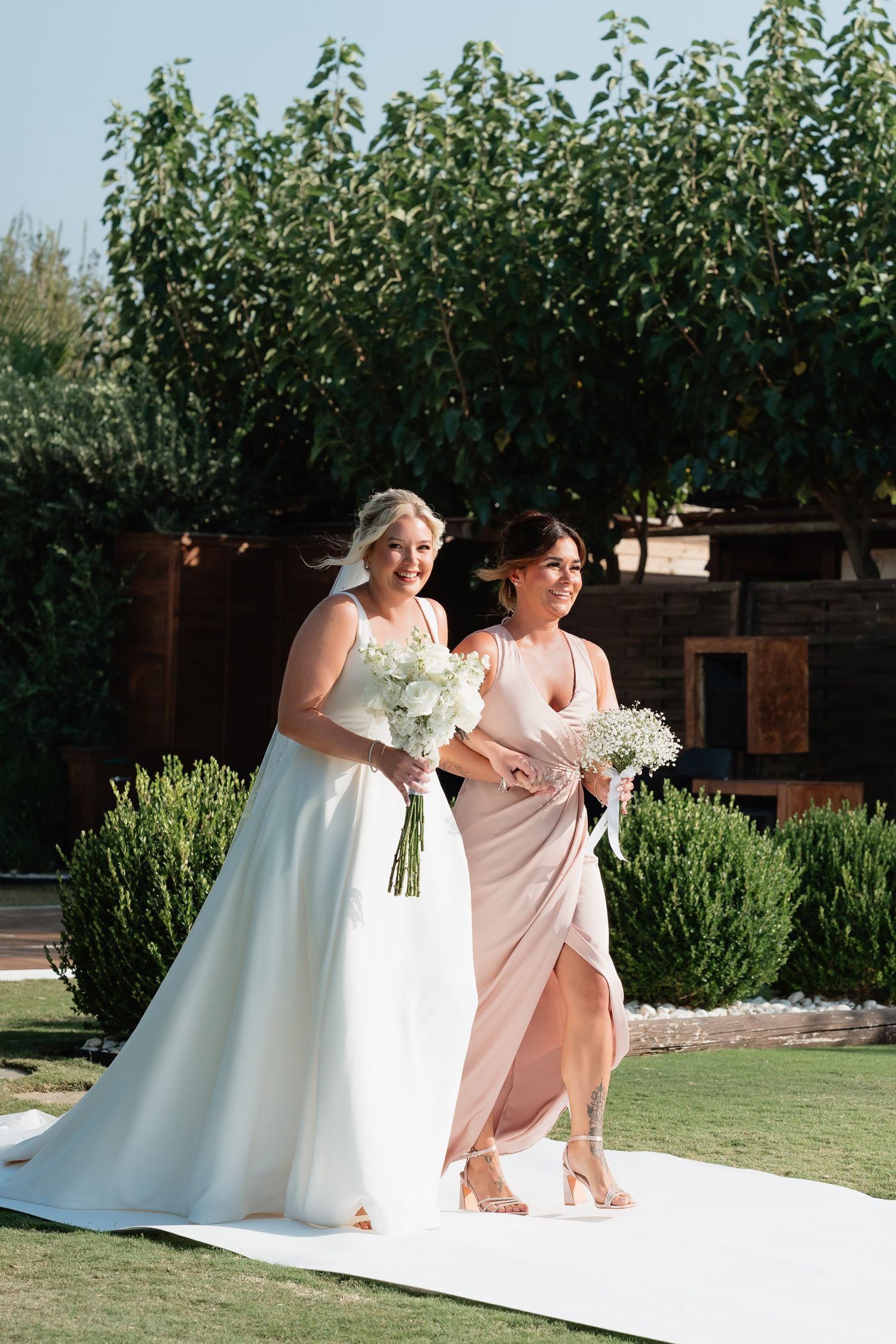 Three bridesmaids in blush pink dresses help the bride adjust her gown in a garden setting during a wedding photoshoot.