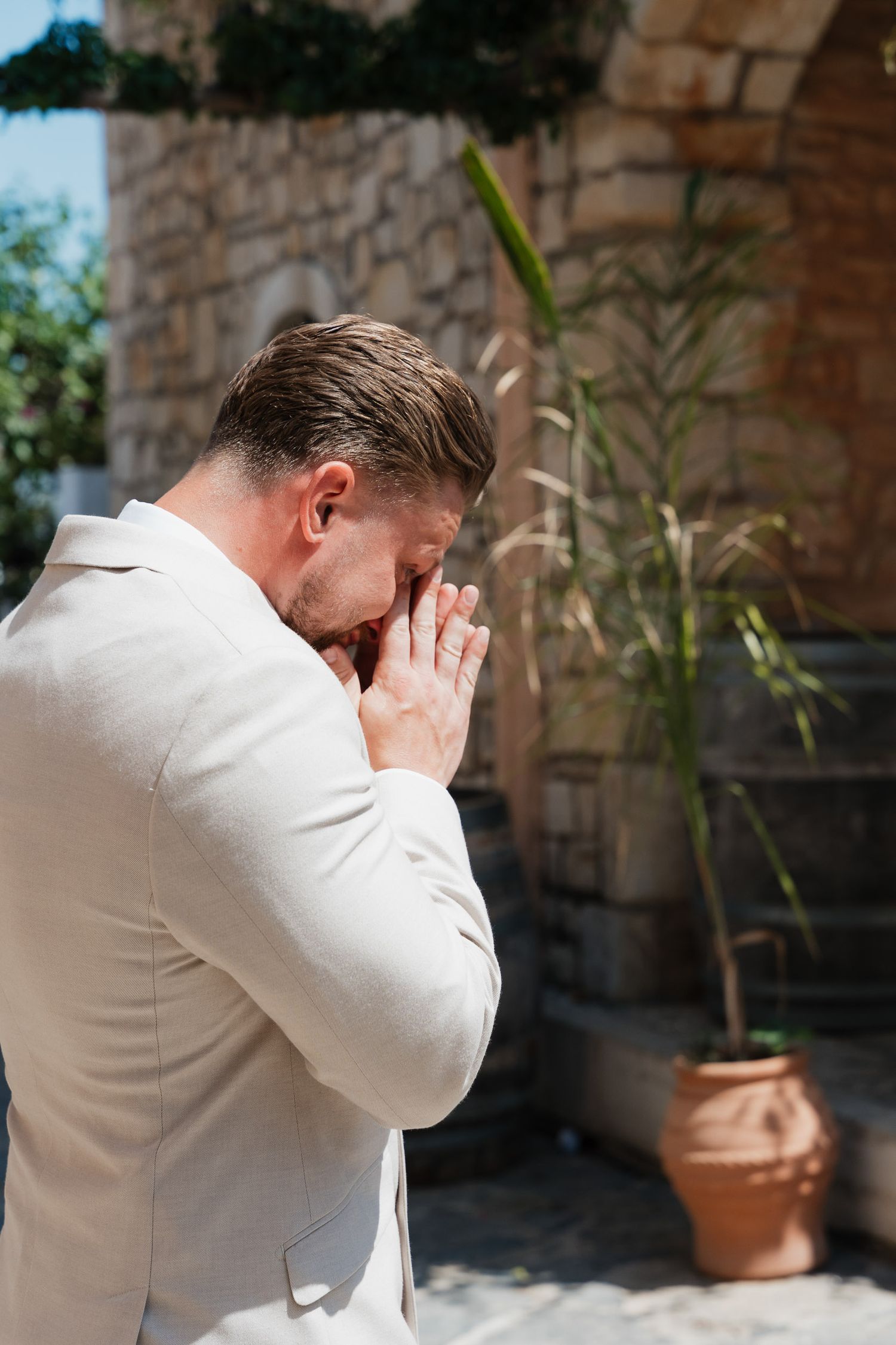 Person in a light beige suit stands in front of a stone wall with potted plants, covering their face with hands.