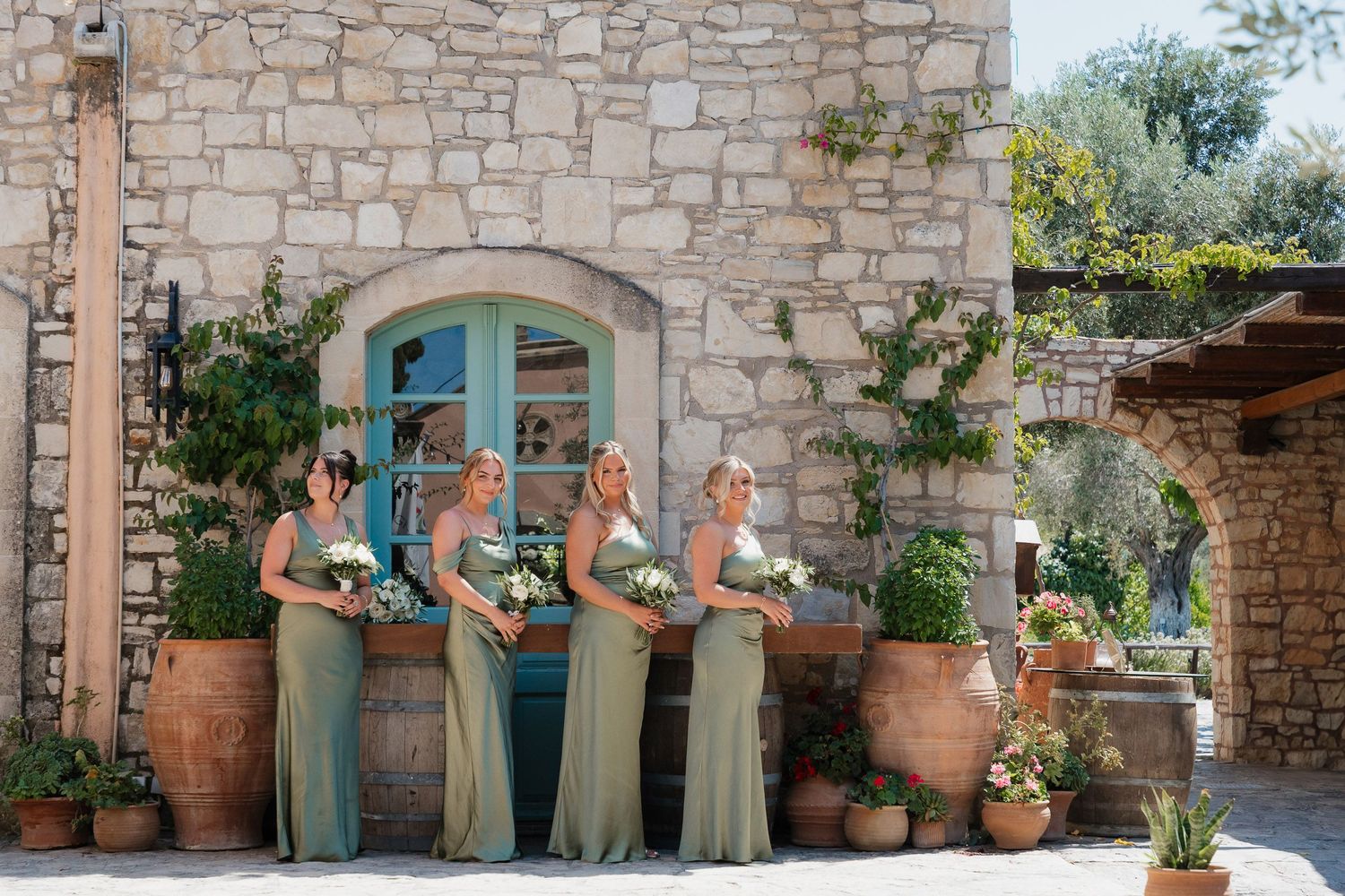 A group of bridesmaids in sage green dresses stand in front of a rustic stone wall with blue door and terracotta planters.