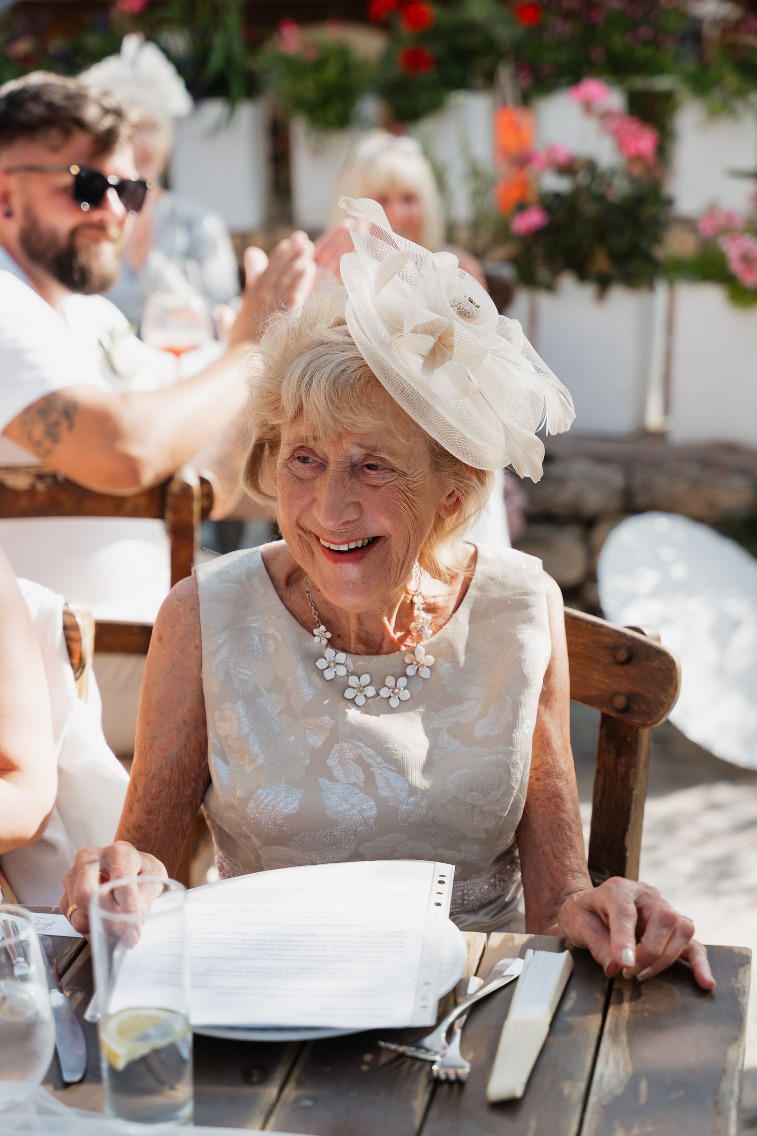 Elegant woman in white lace dress and pearl necklace smiling while seated at outdoor dining table.