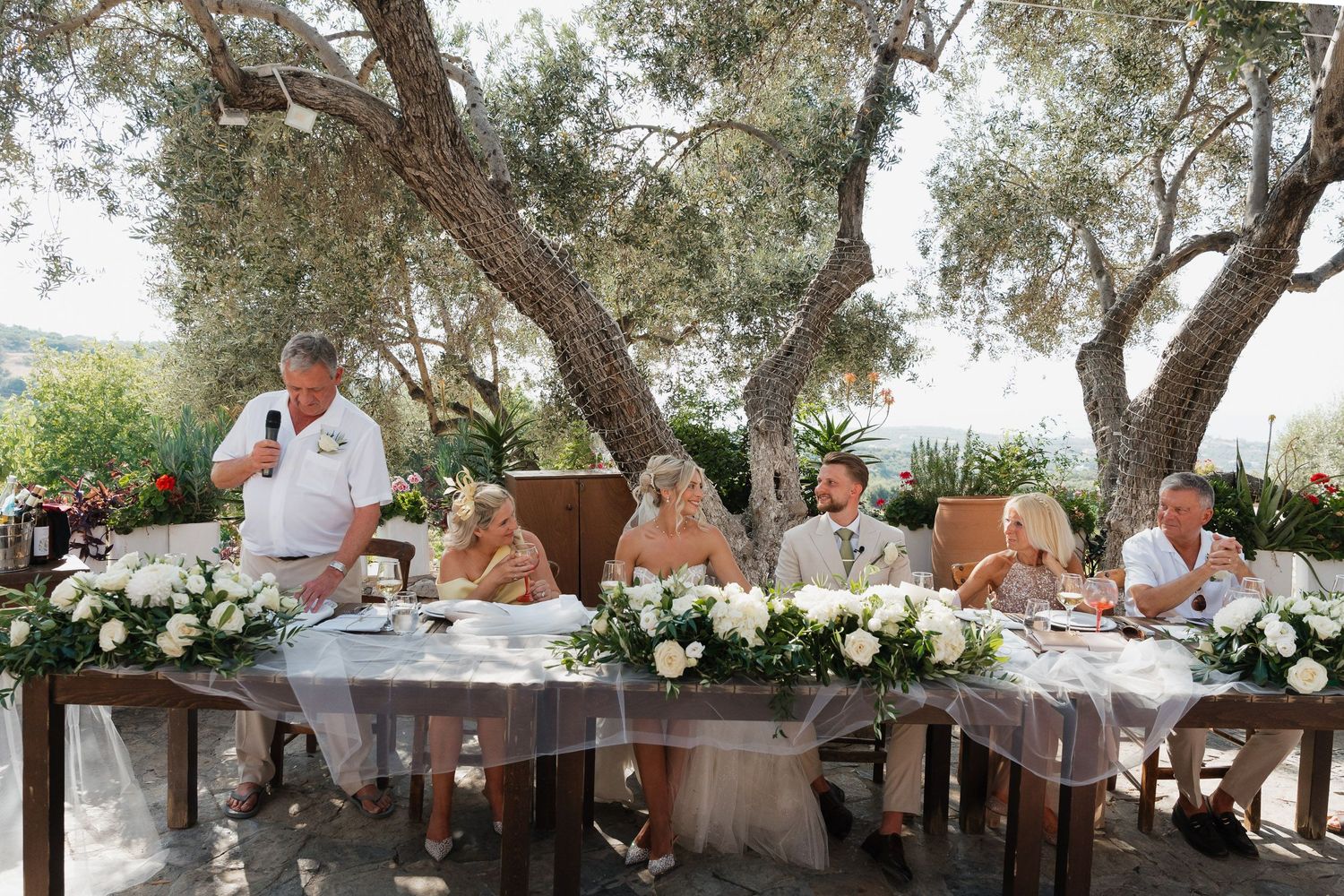 Wedding guests enjoy an elegant outdoor reception dinner under olive trees at a Mediterranean venue.