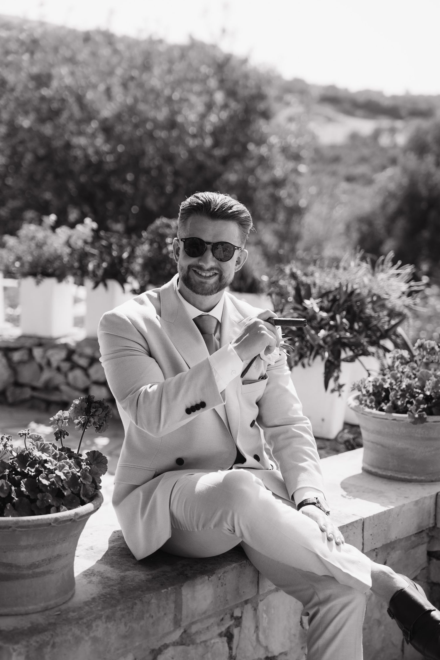 A person in a white suit and sunglasses sits casually on a stone wall surrounded by potted plants in a garden setting.