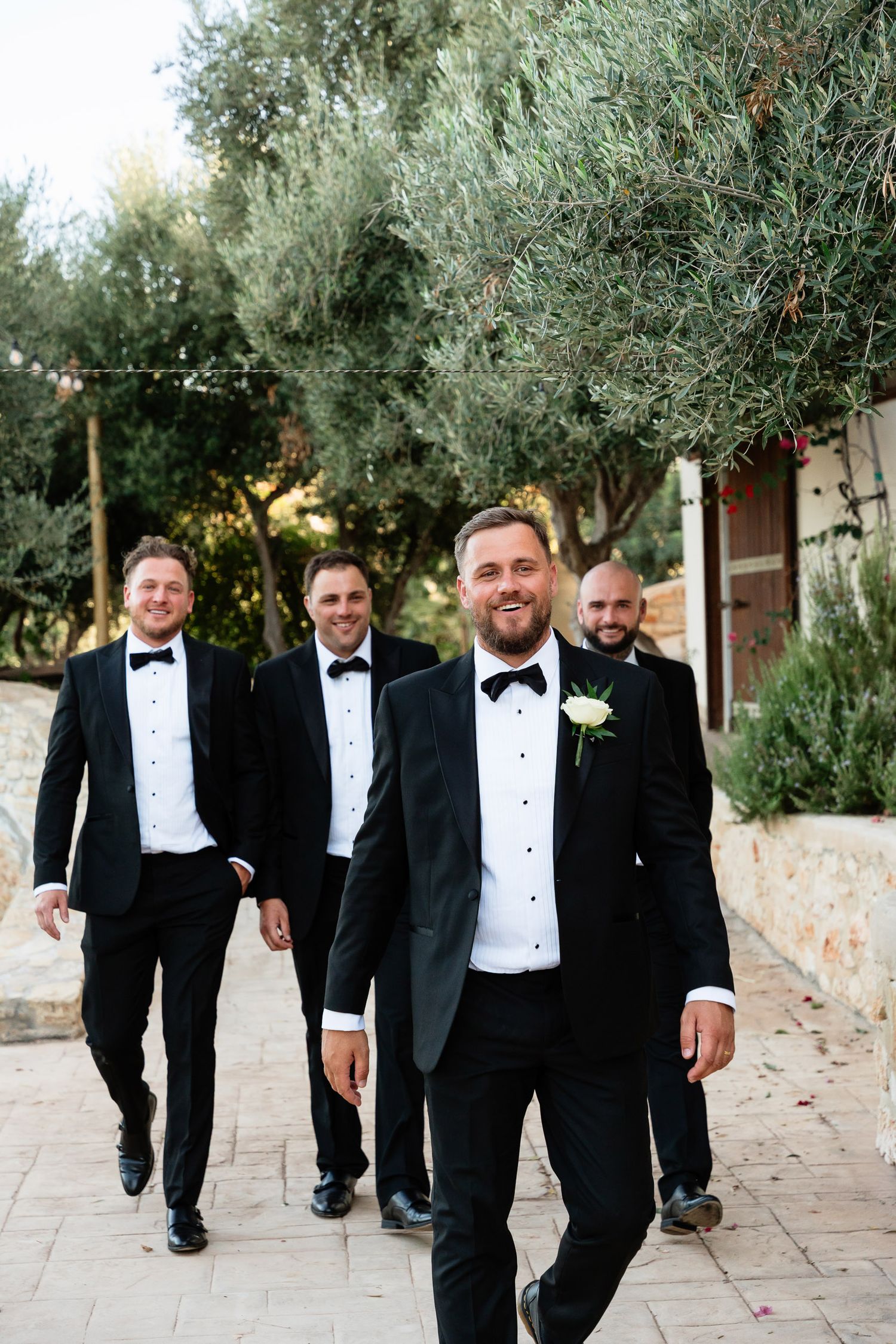 Groomsmen in black tuxedos and bow ties walk together along a stone path surrounded by olive trees.