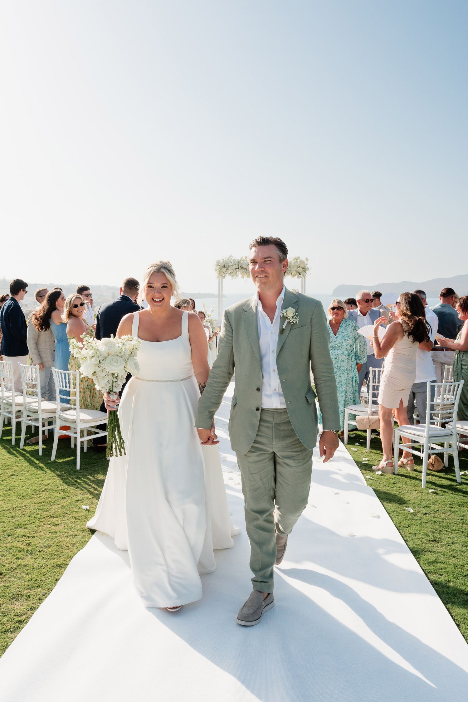 A couple walks down an outdoor wedding aisle with white chairs and ocean views in the background.