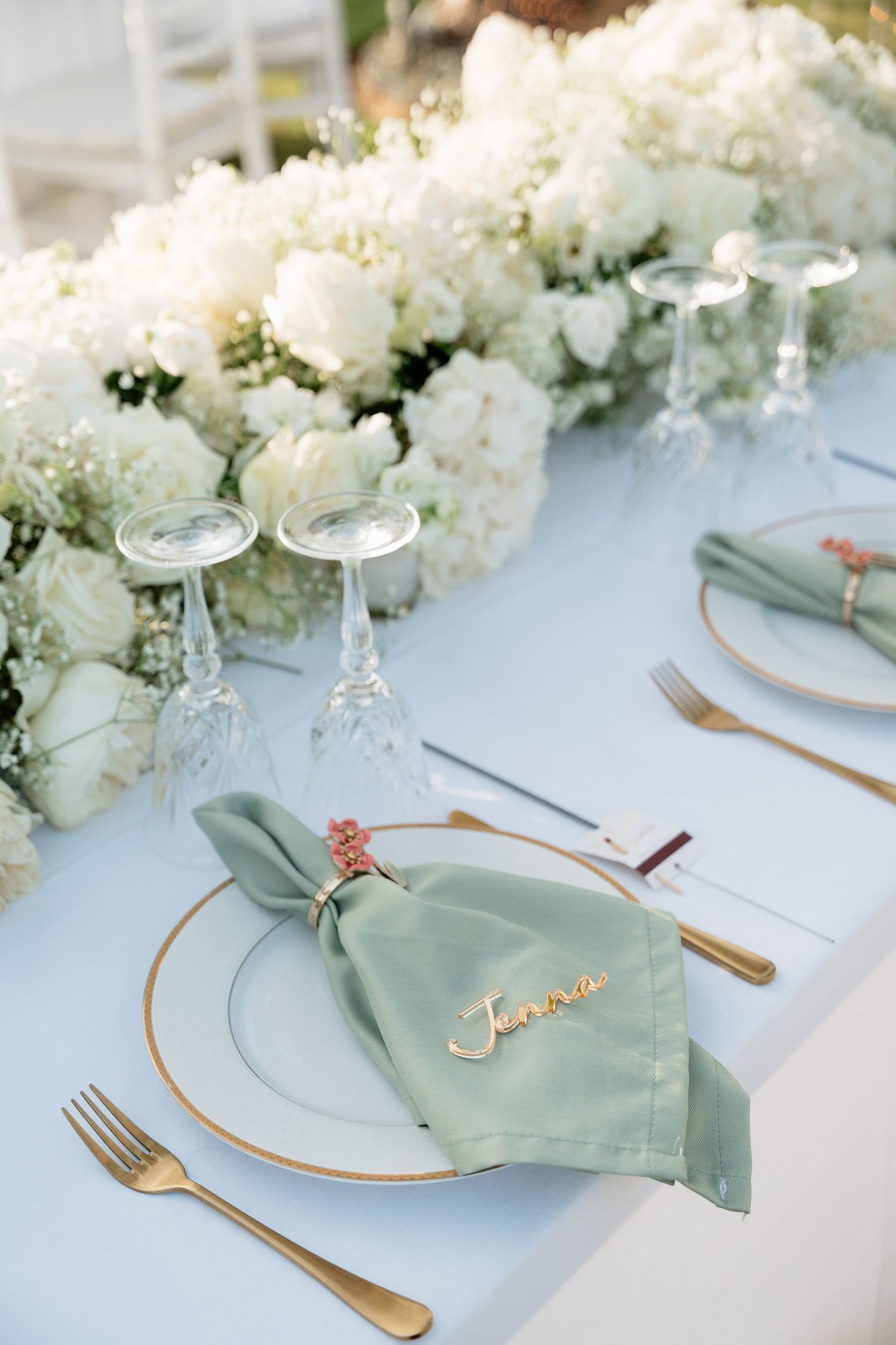 Elegant wedding table setting with white floral centerpieces, sage green napkins, and gold-rimmed plates and cutlery.