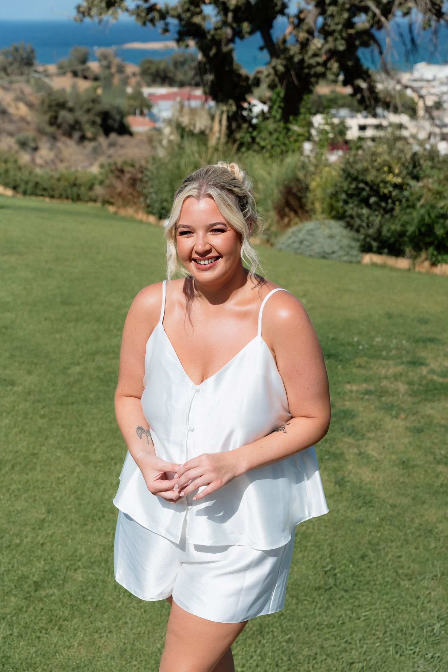 A person in a white summer outfit stands on a grassy hill overlooking the ocean on a sunny day.