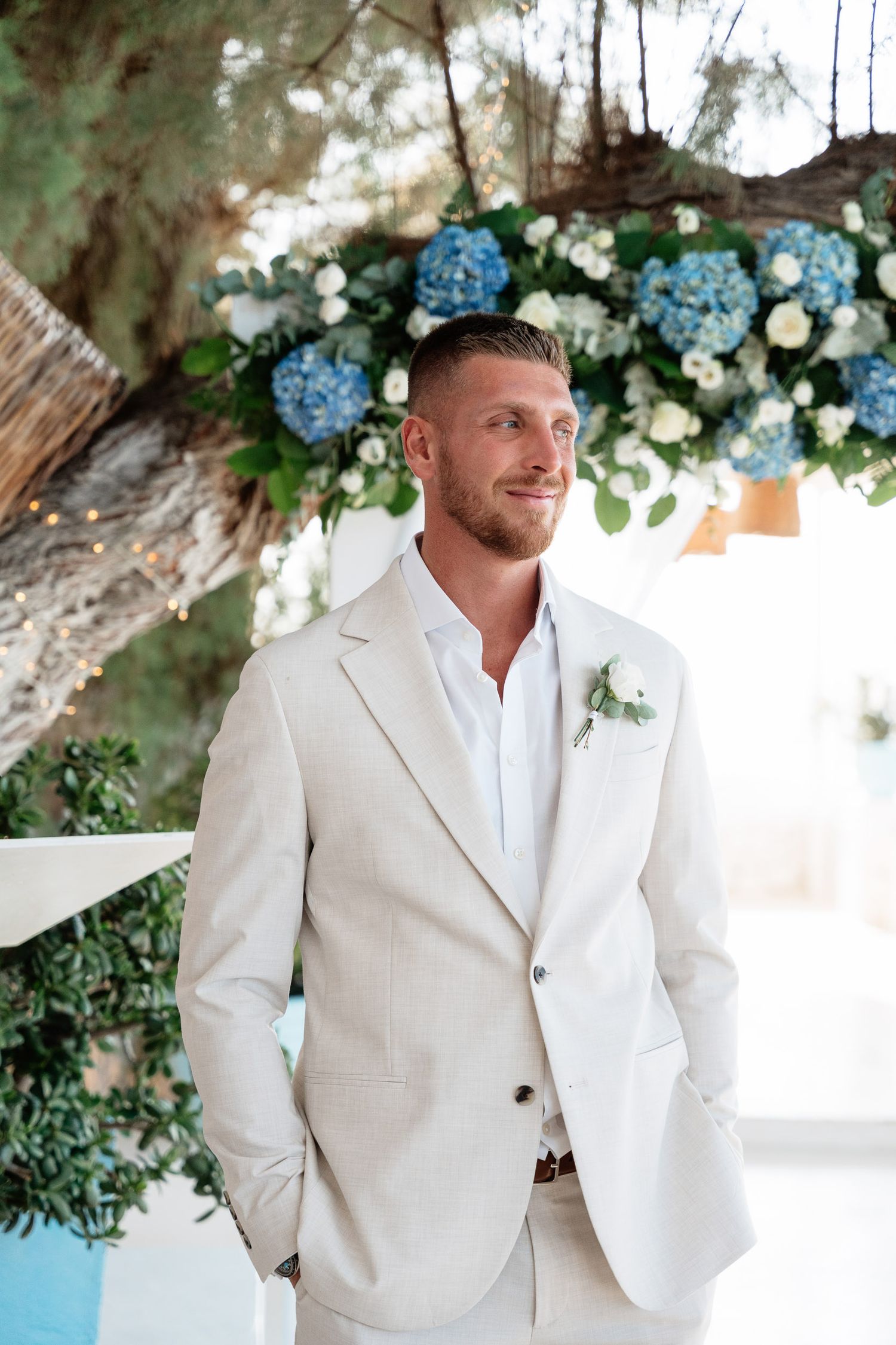 A groom in a light beige suit and white shirt stands under a floral arch with blue hydrangeas and white flowers.