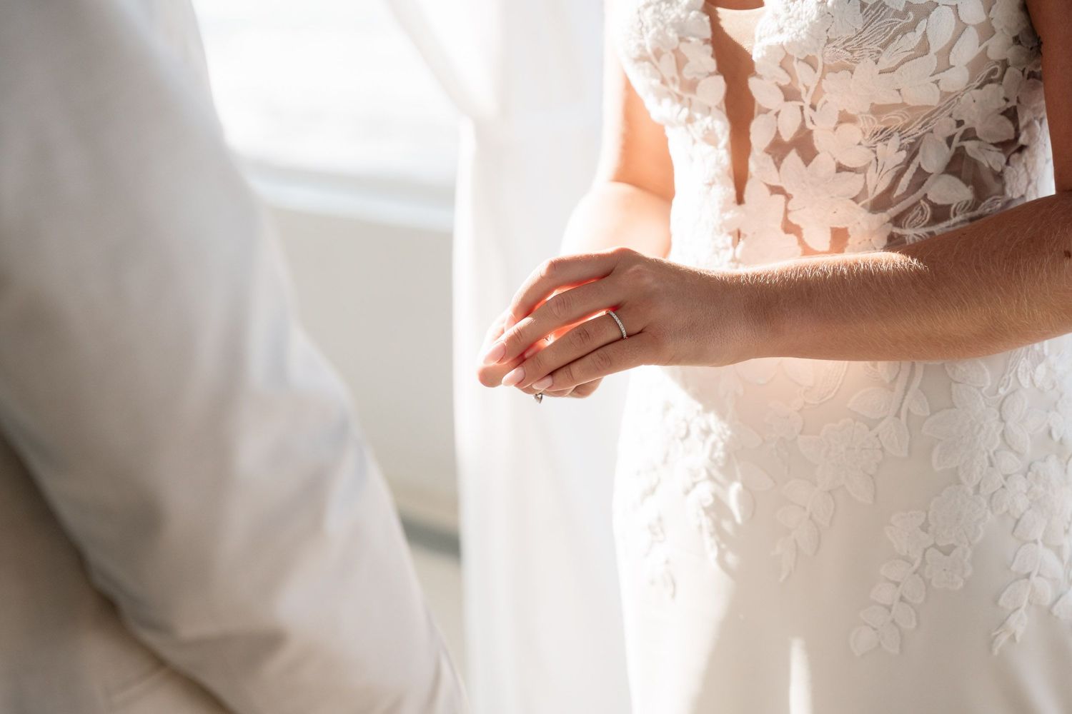 Close-up detail of delicate lace wedding dress and hands in soft natural window light.