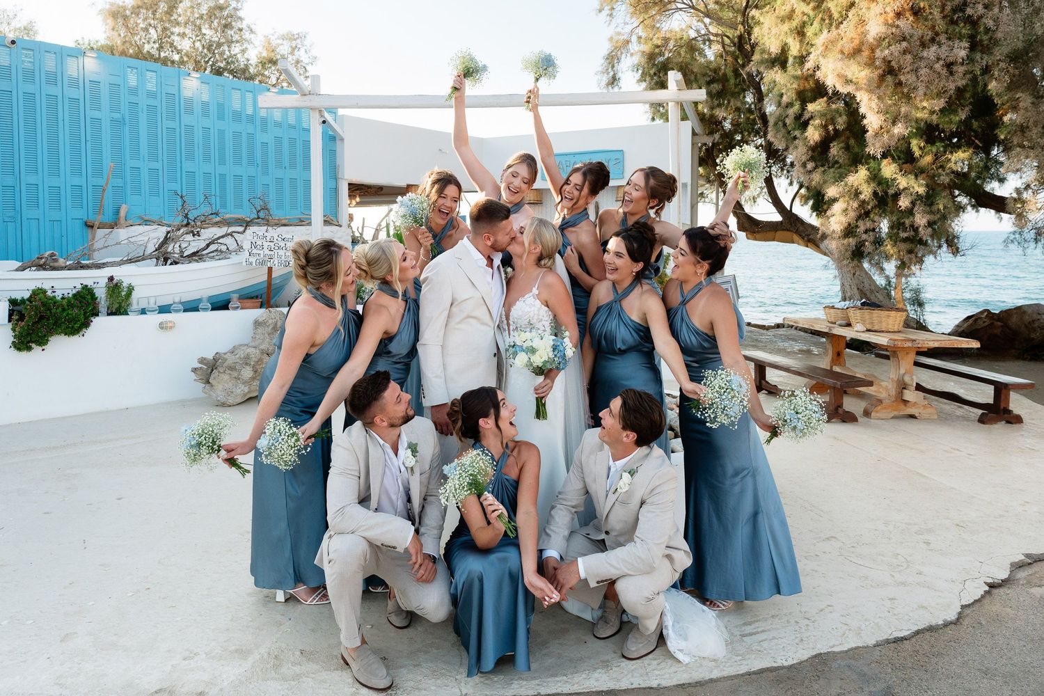 Wedding party sits together on white stone steps overlooking a Mediterranean coastal view at sunset.