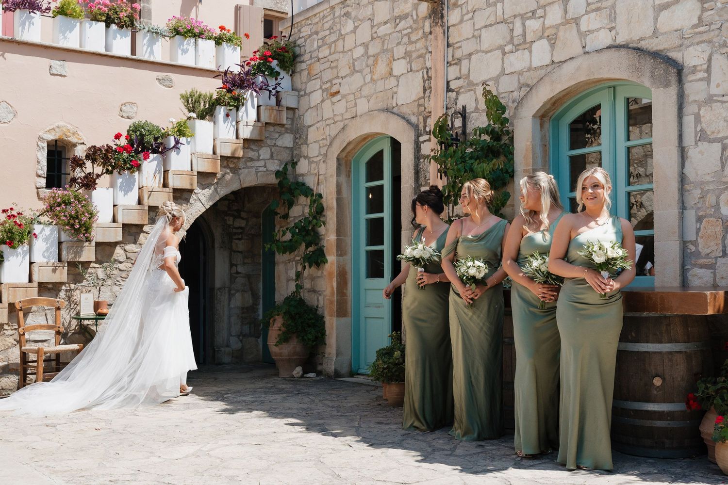 A wedding party gathers in front of a rustic stone wall with turquoise doors and hanging flower baskets.