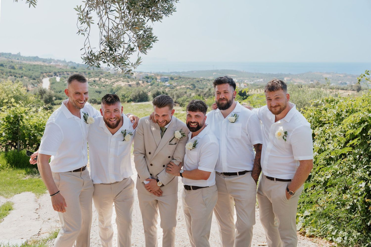 A group of groomsmen in light colored suits pose together on a scenic hillside path surrounded by greenery.