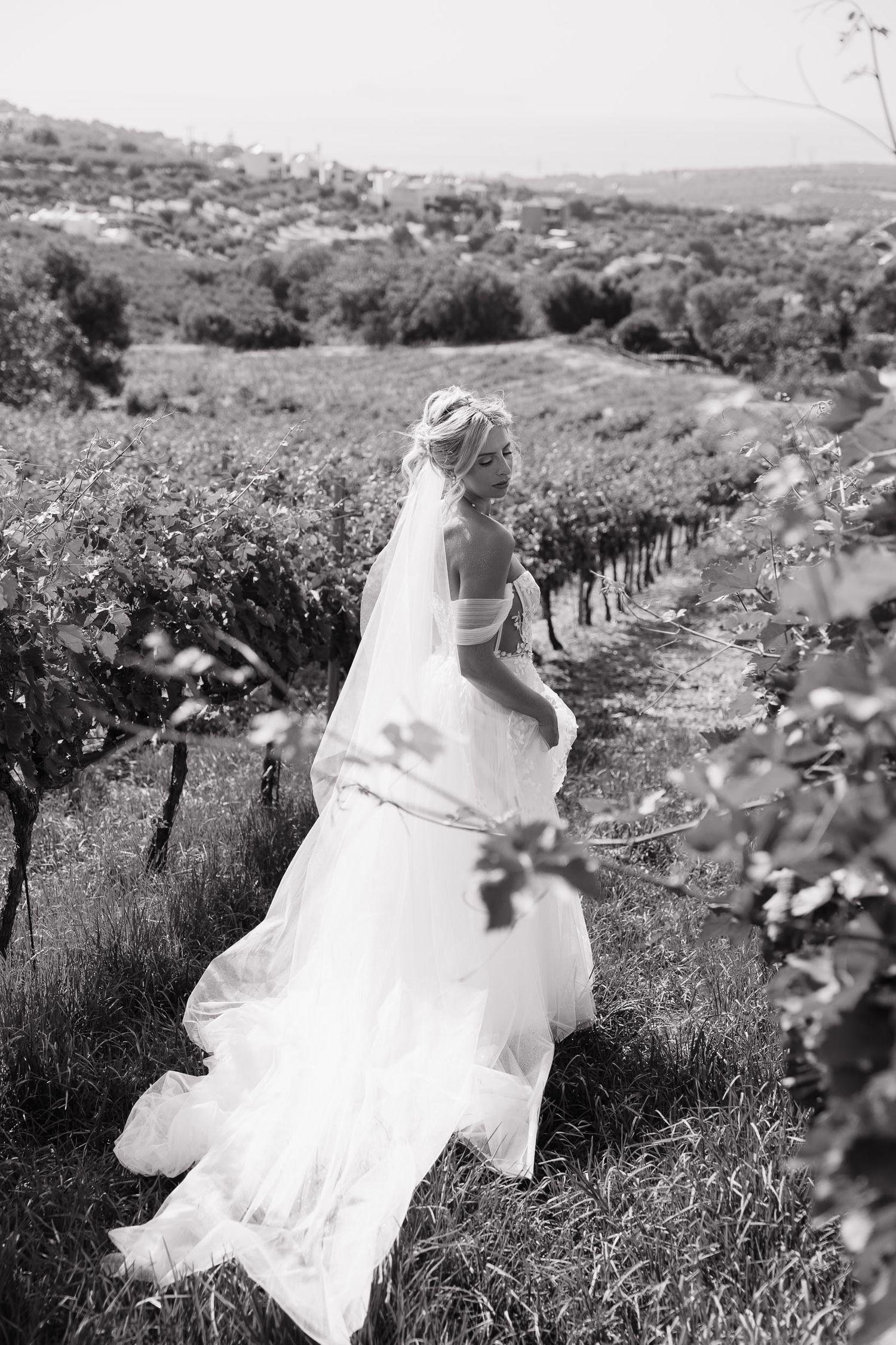 A figure in a flowing white wedding dress runs through a vineyard landscape in black and white.