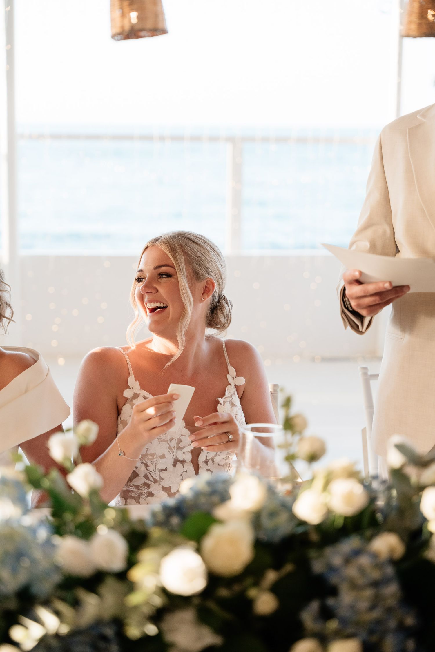 A joyful moment during wedding speeches at an elegant reception with floral centerpieces and soft lighting.