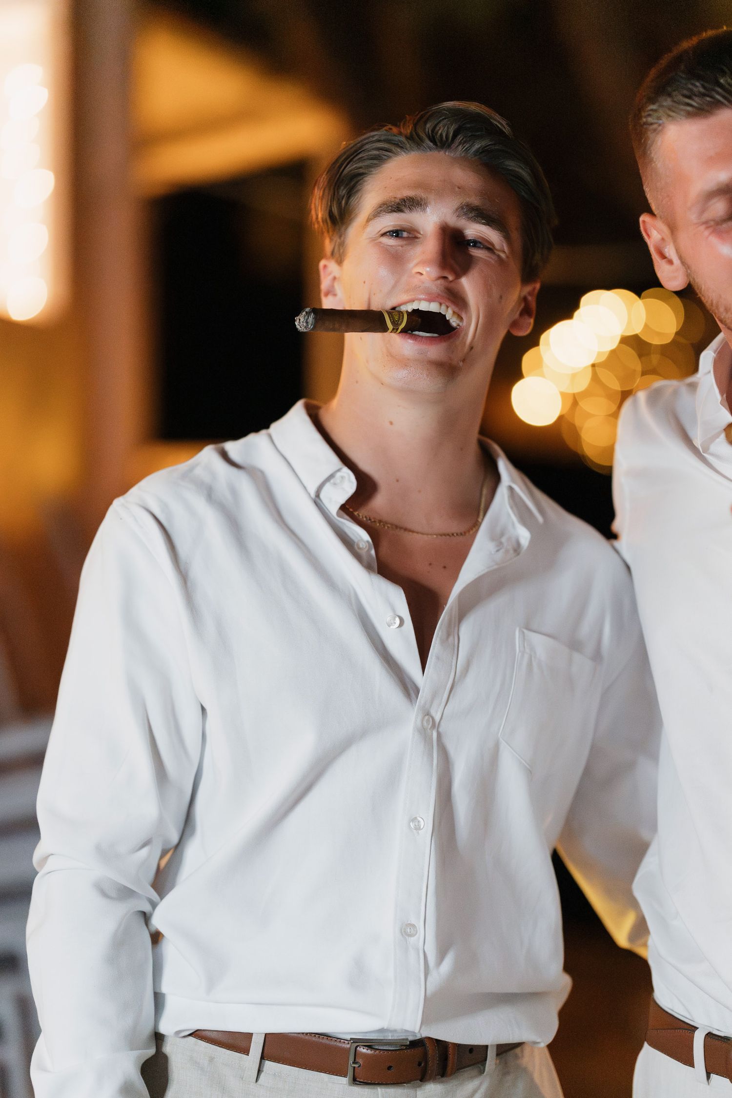 A person in a crisp white button-down shirt and brown belt laughs joyfully in an indoor setting with bokeh lights behind them.