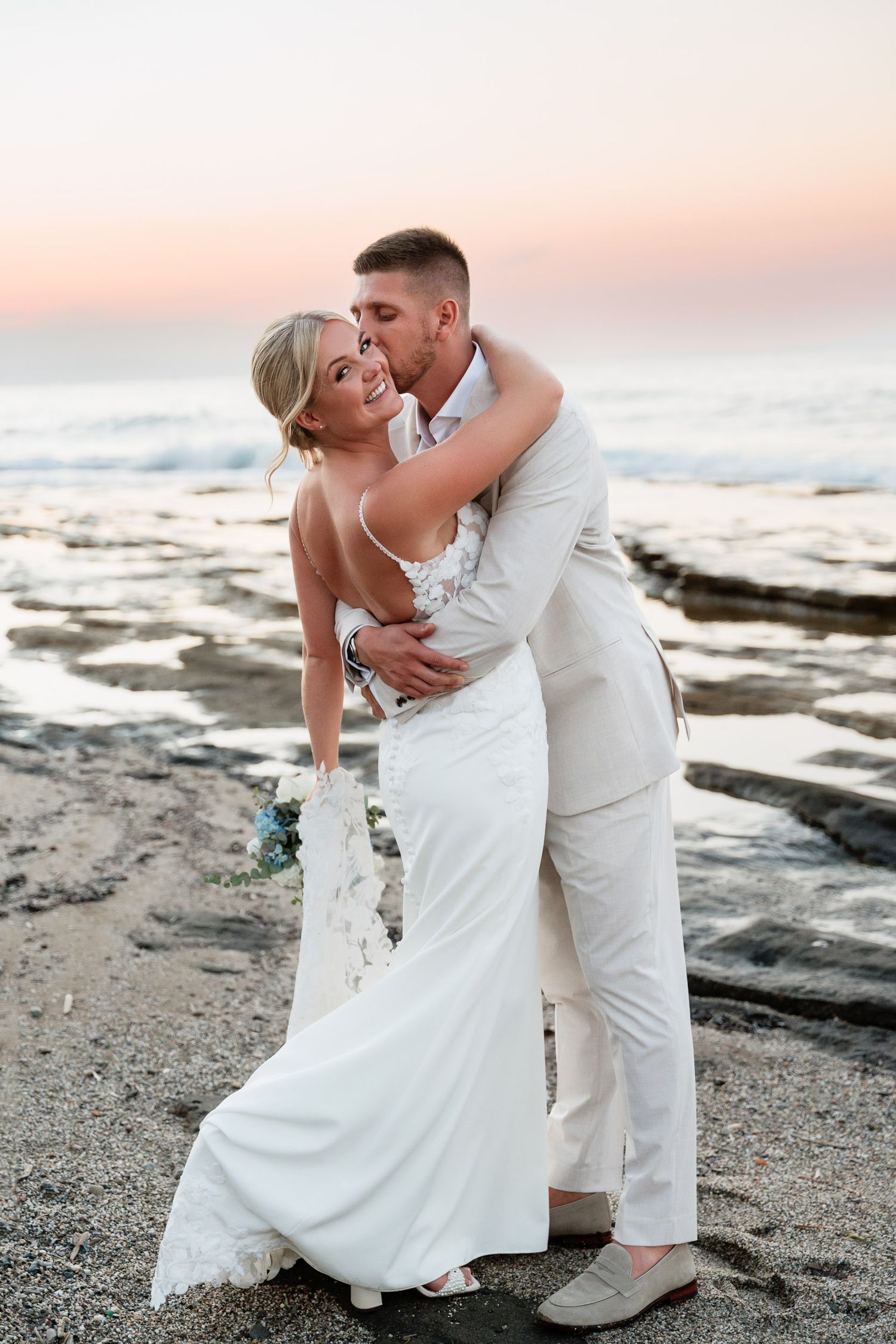 A romantic beach wedding couple embraces during sunset on a rocky shoreline with soft pink sky in the background.