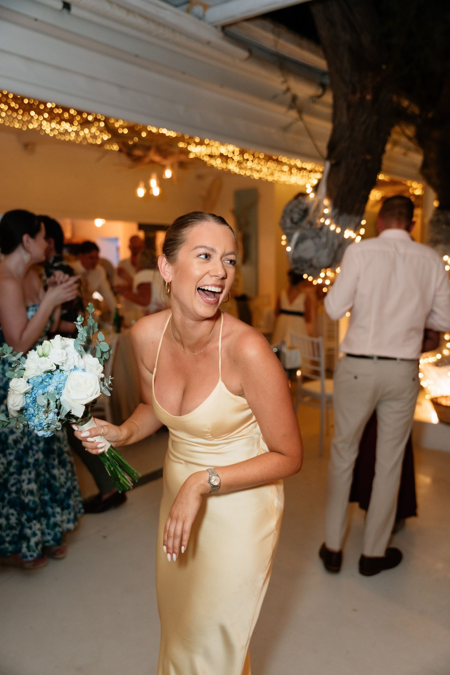 A person in a yellow spaghetti strap dress dances and smiles on a wedding reception dance floor with string lights above.