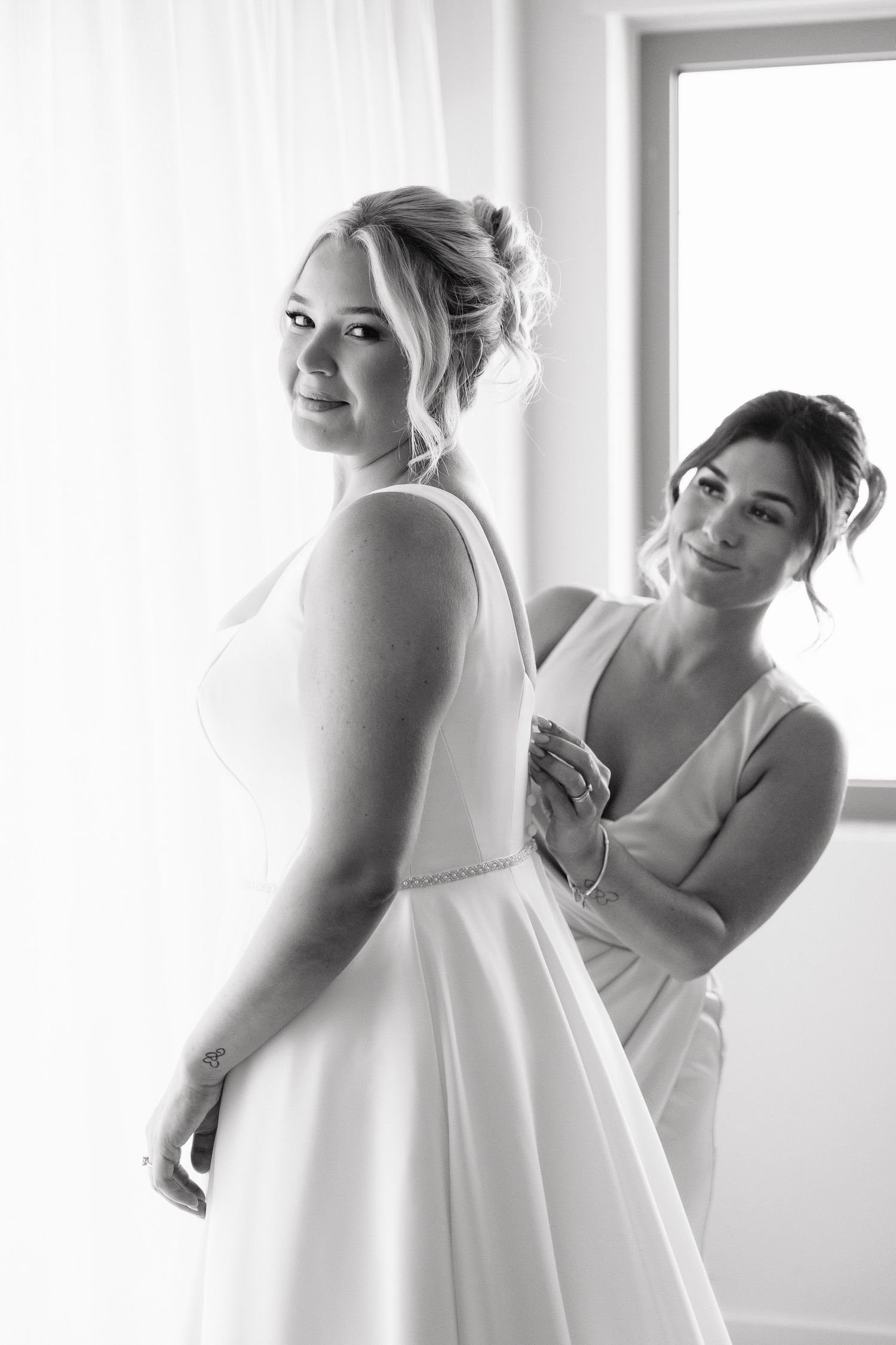 Black and white photo series showing a bride getting ready in a sleeveless white wedding gown by a bright window.