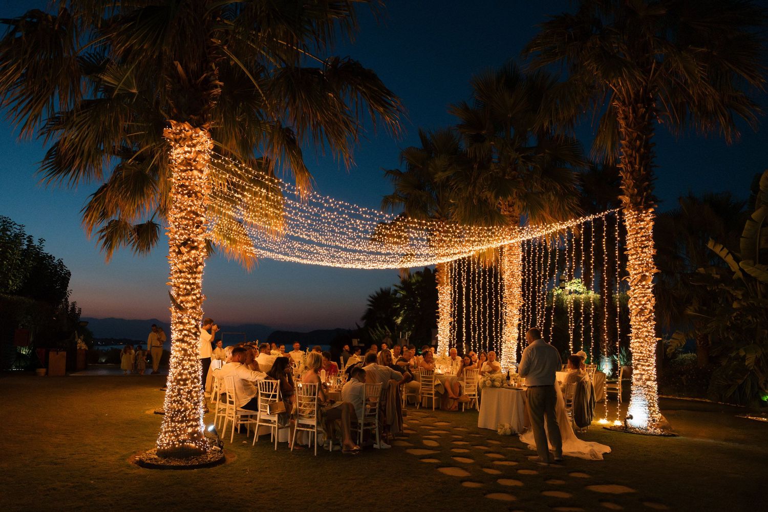 String lights illuminate palm trees and create a romantic outdoor dining atmosphere at dusk.