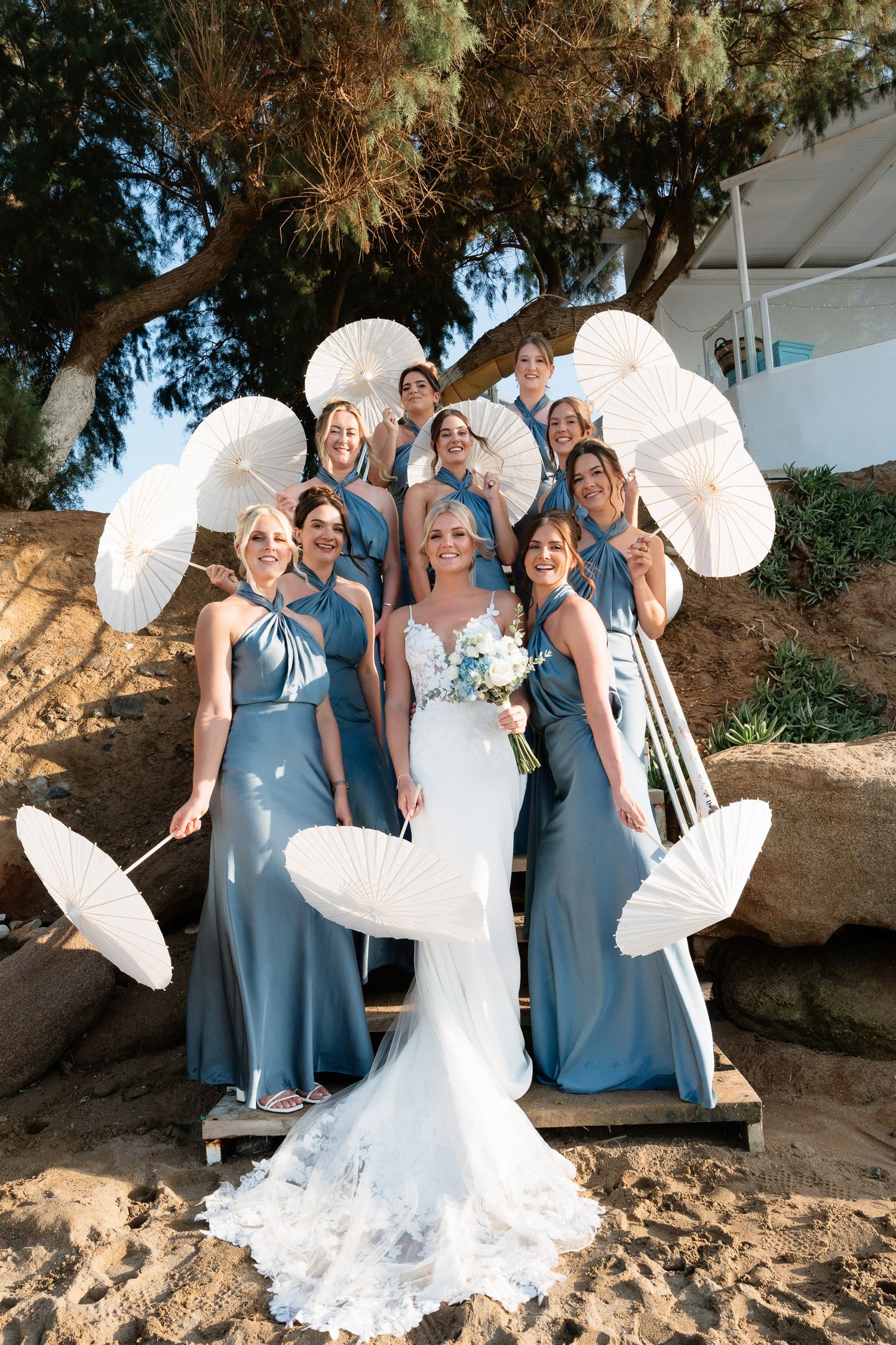A bridal party in dusty blue dresses poses with white parasols on a beach during a sunny wedding photoshoot.